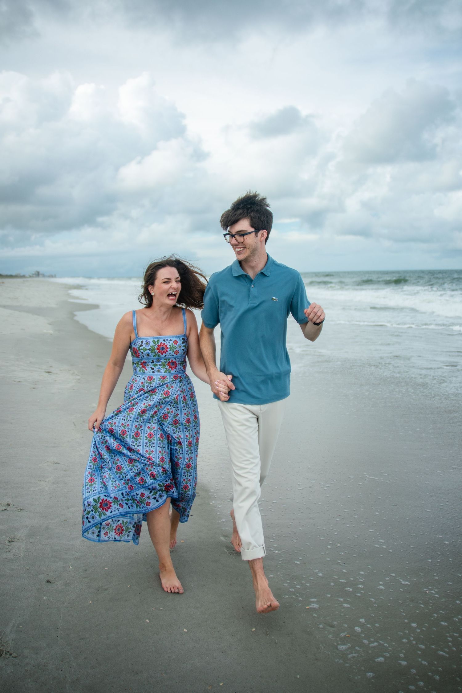 A playful couple runs and laughs together along the shoreline of a cloudy beach.