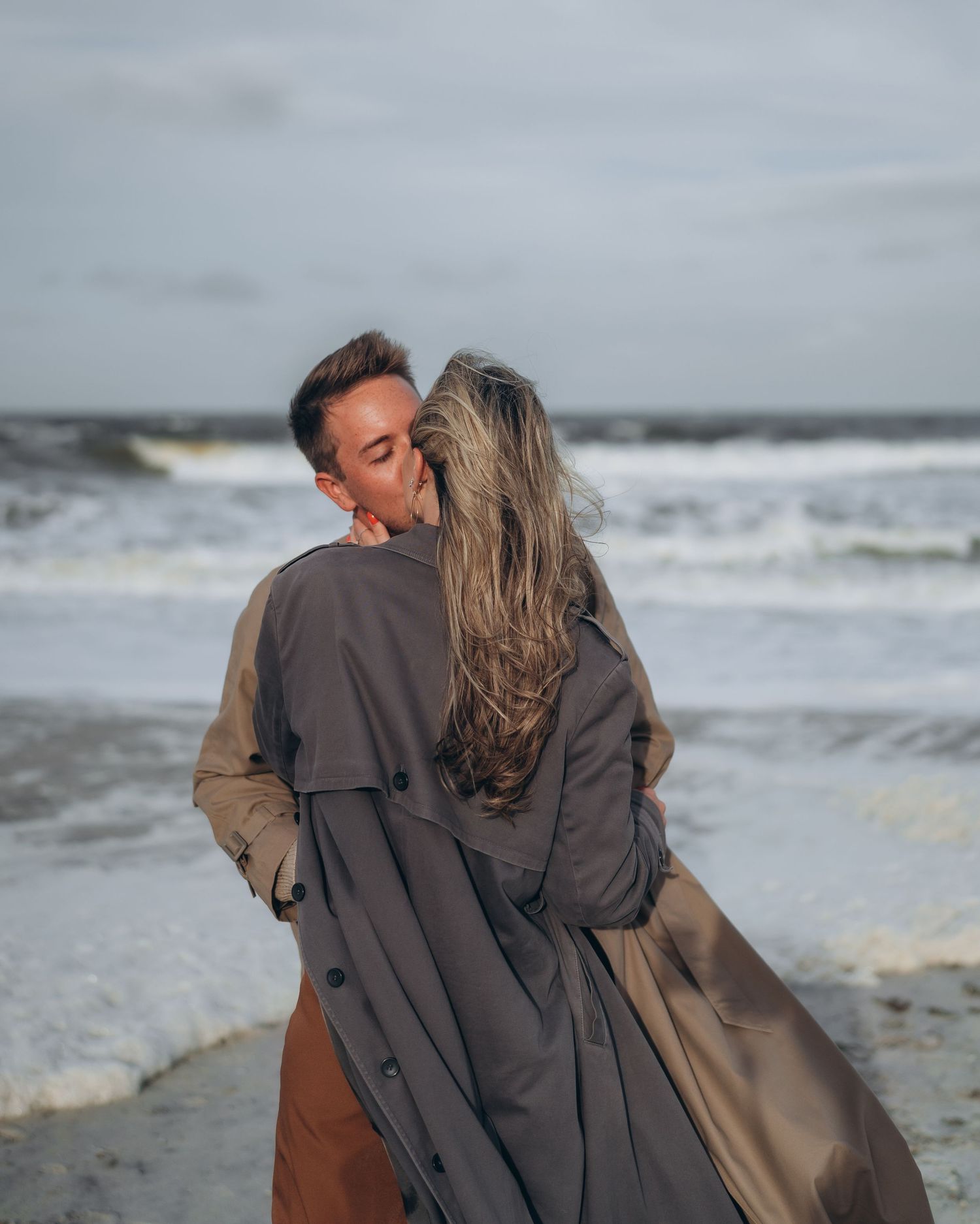 A couple in dark coats share a romantic moment on a windy beach with crashing waves behind them.