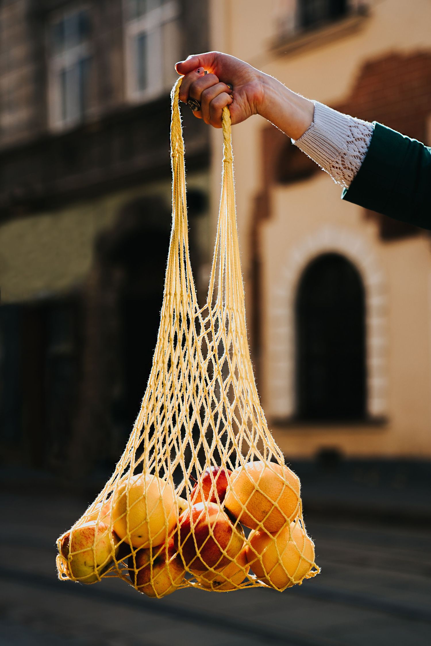 Hand holding a yellow mesh string bag filled with fresh oranges and fruits on a blurred urban street background.
