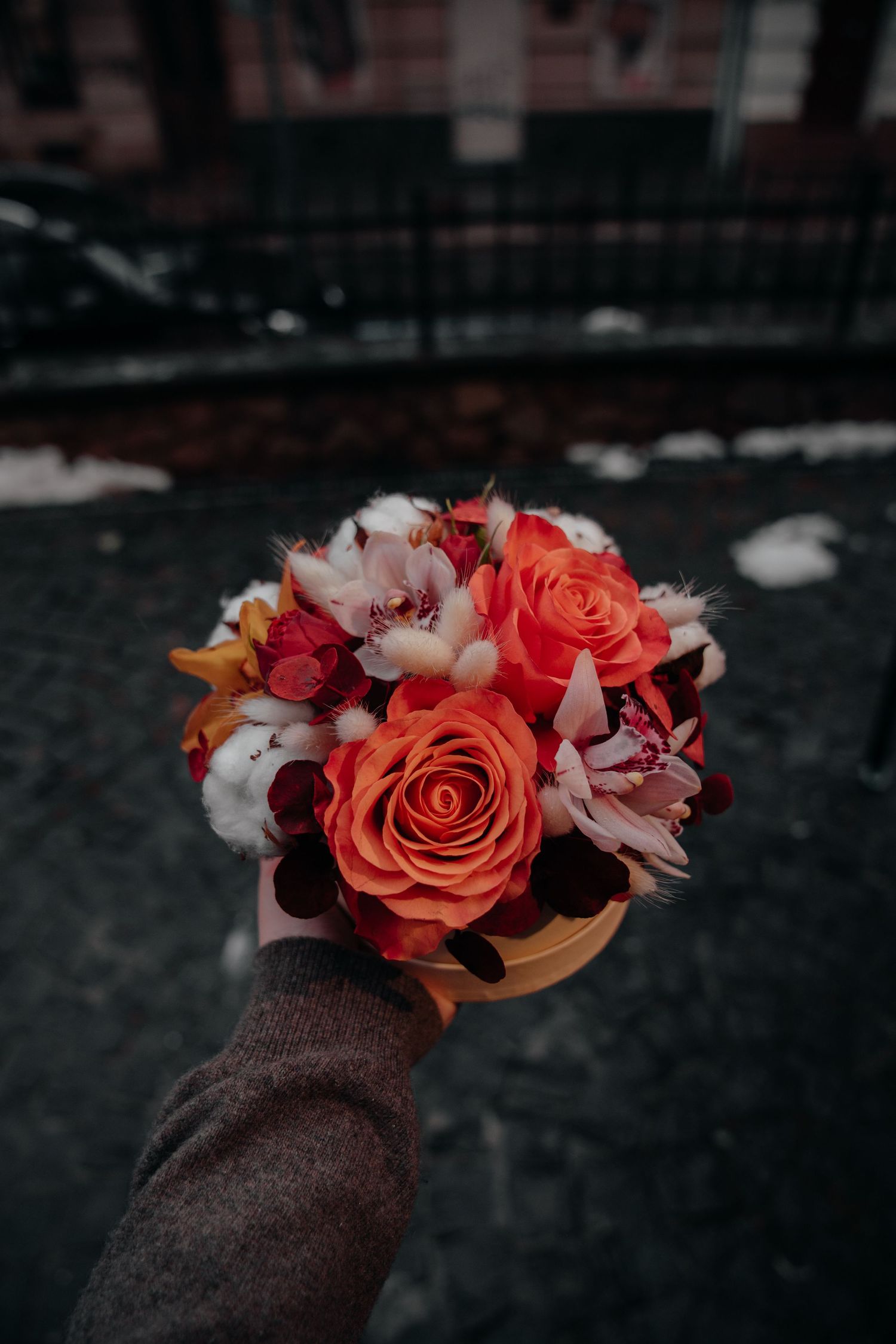 Hand holding a golden box bouquet with orange roses, red flowers, and white cotton blooms against a dark urban background.