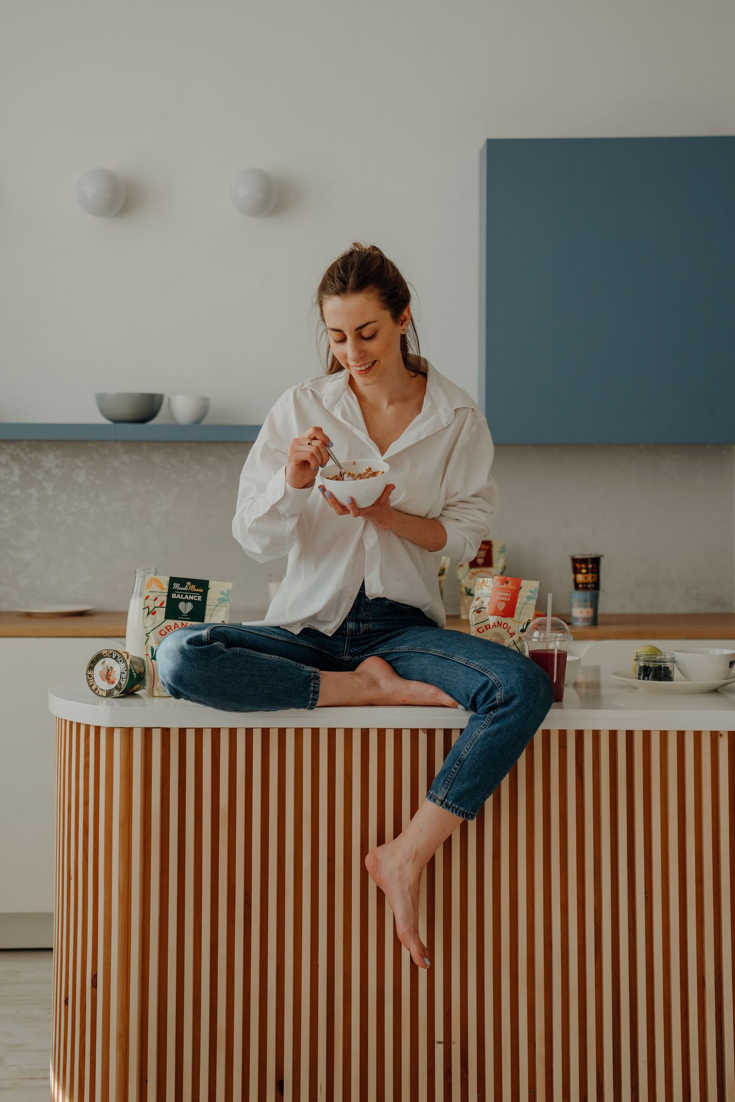 Woman in white shirt sitting cross-legged on kitchen counter eating granola from a white bowl.