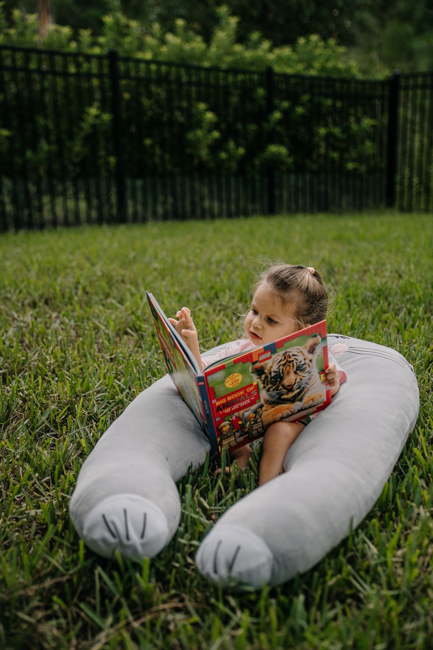 Toddler girl reading a colorful book outdoors while resting on a large gray body pillow on green grass.