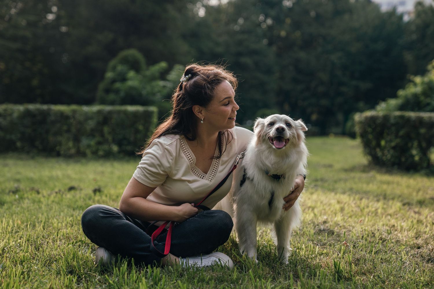 Woman sitting on grass with her happy white fluffy dog in a lush green park on a sunny day.