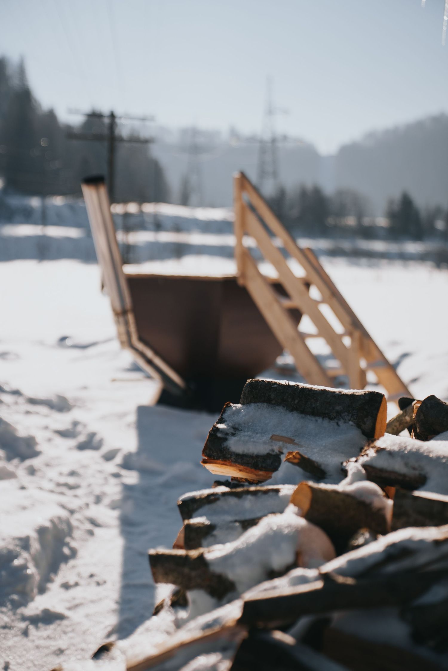 Snow-covered firewood pile in winter landscape with wooden stairs and blurred background.