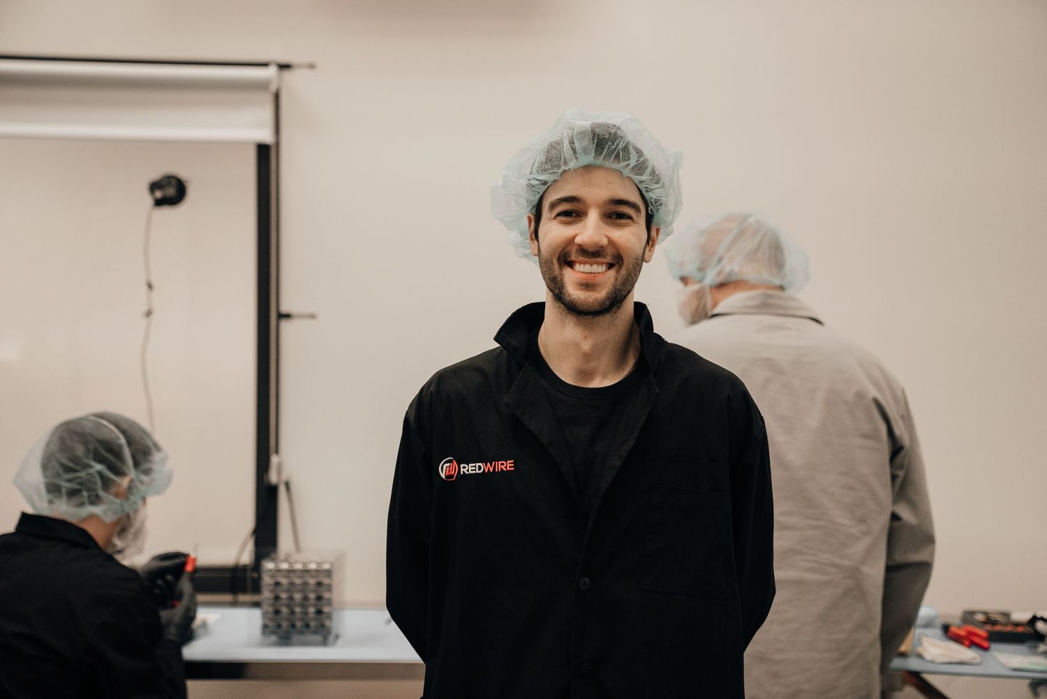 Smiling young male scientist in hairnet and black lab coat stands confidently in a professional laboratory setting.