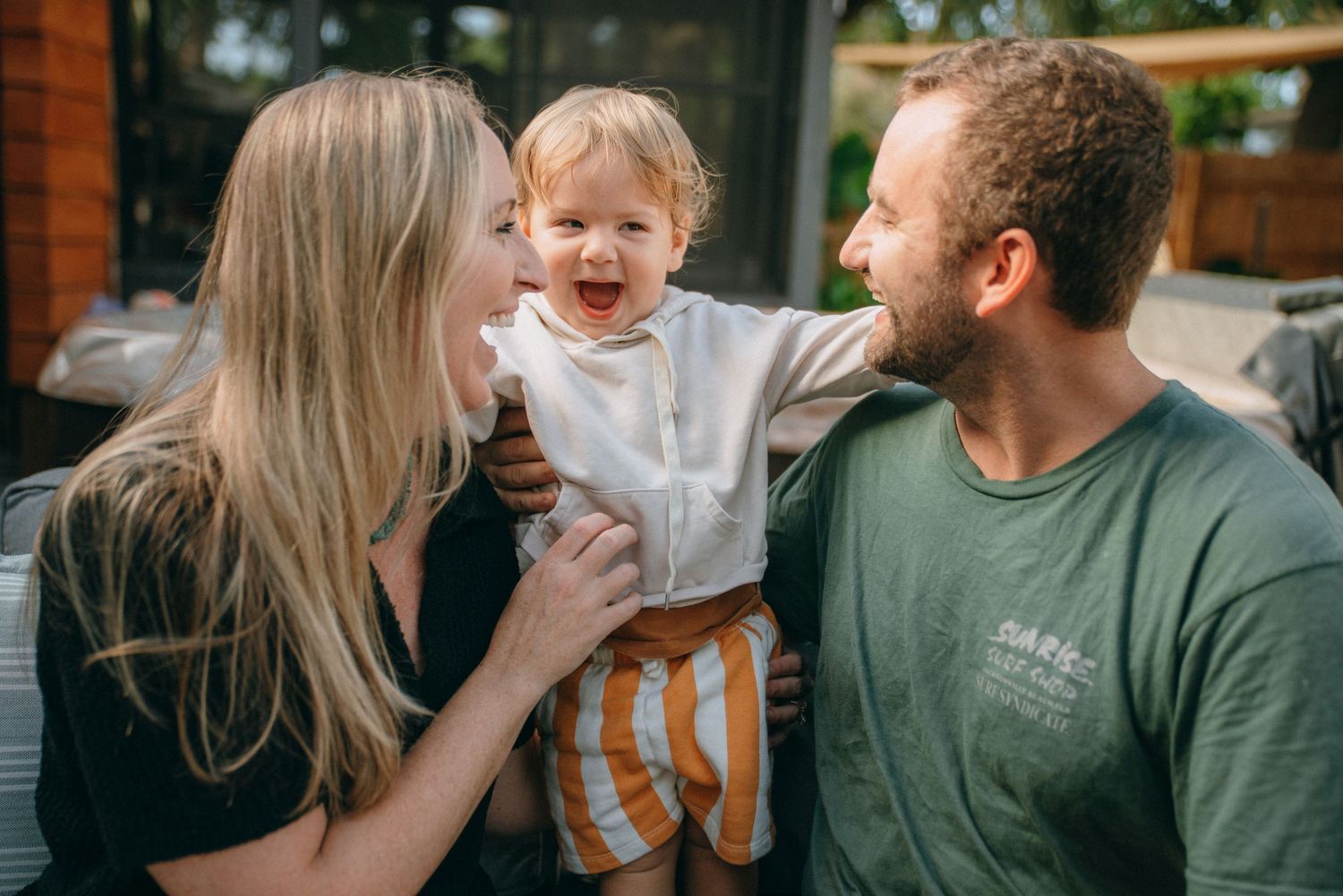 Family laughing and playing together outdoors near a wooden fence.