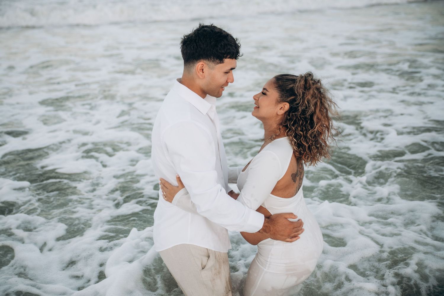 Romantic couple embraces while standing in ocean waves at sunset.