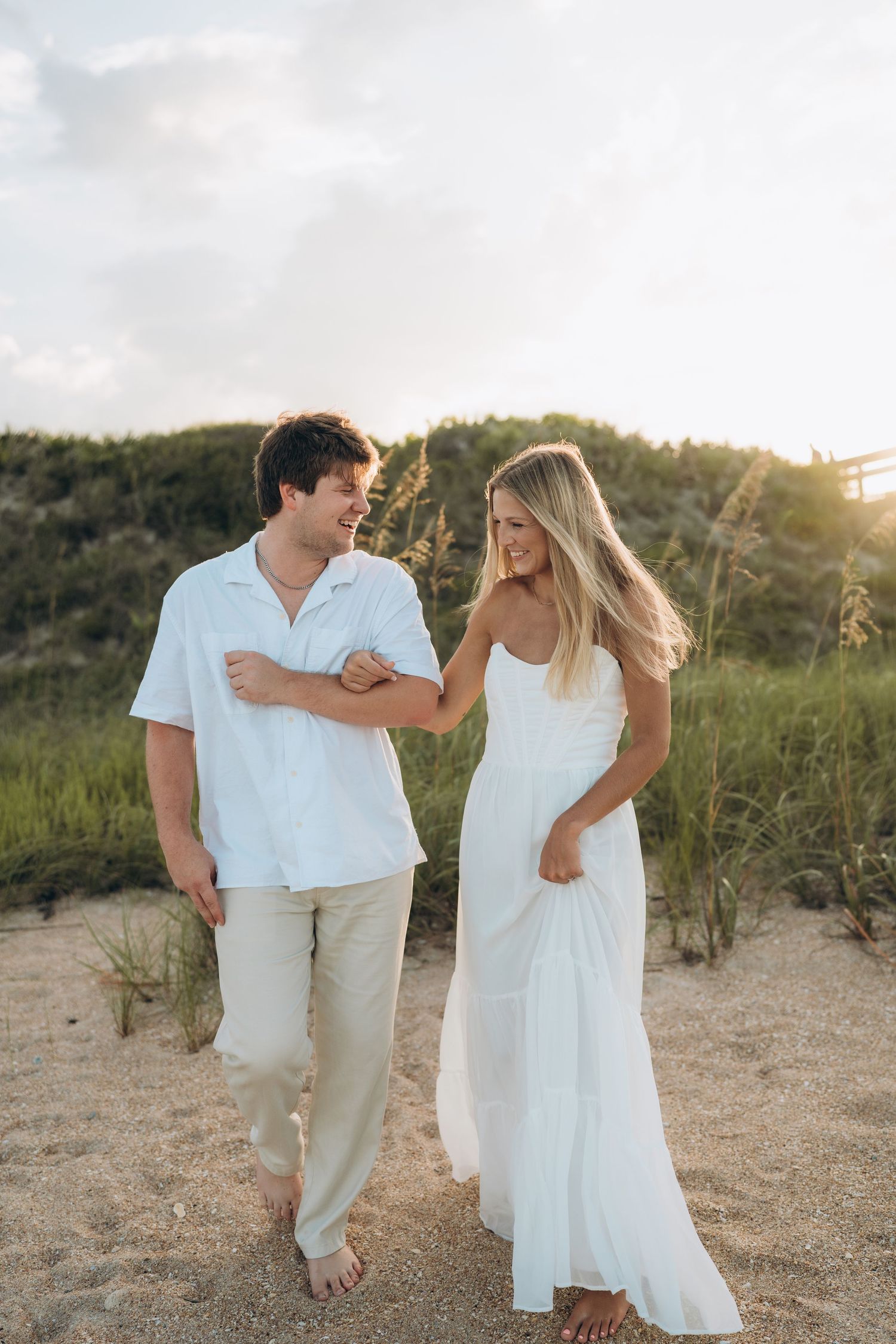 A playful couple in white clothing dances together on a sandy beach path.