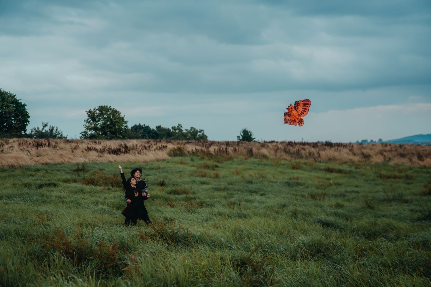 Person in dark clothing with red balloon in grassy field under cloudy sky.