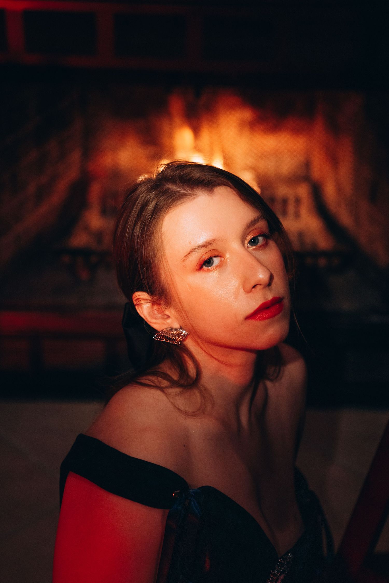 Young woman with red lips and earrings poses elegantly in front of a glowing fireplace in an off-shoulder dress.