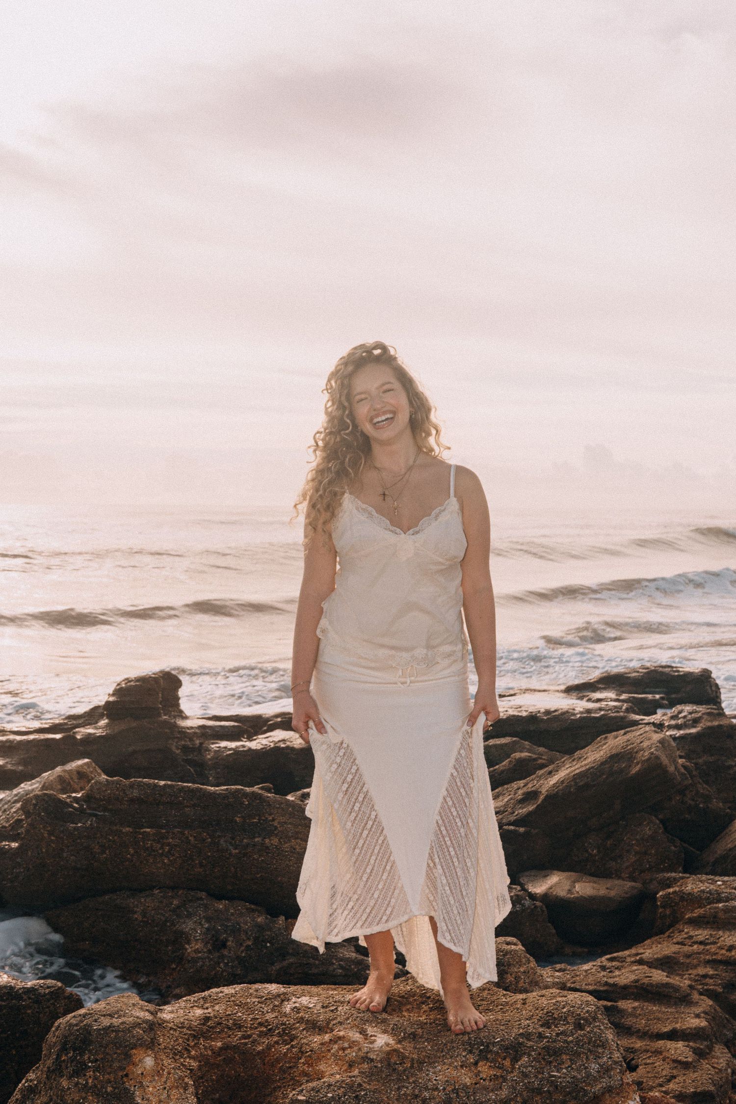A woman in a white dress laughs joyfully while standing barefoot on rocky shore with ocean waves behind her.