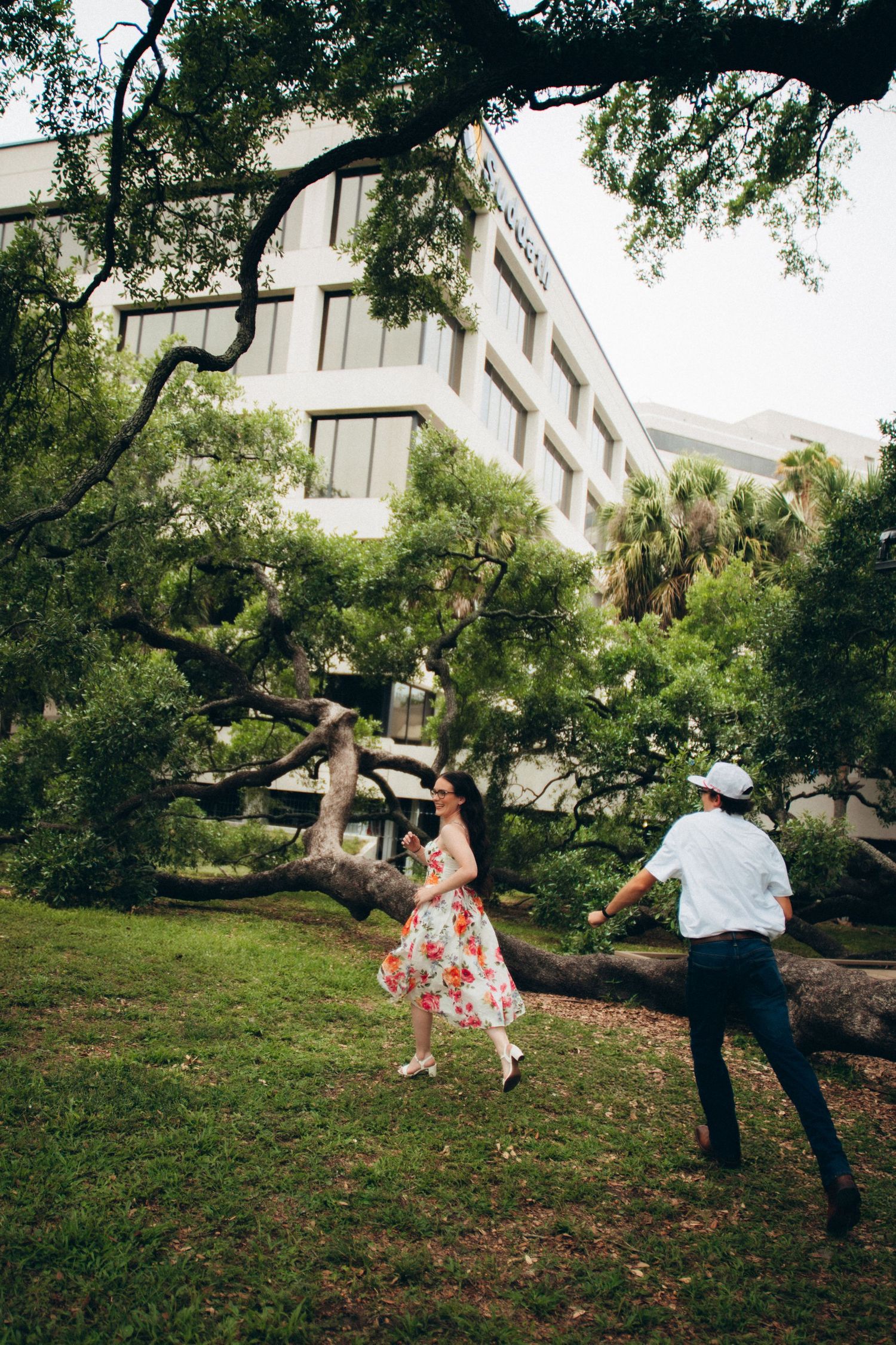 Couple dancing playfully under large oak tree branches with building visible in background.