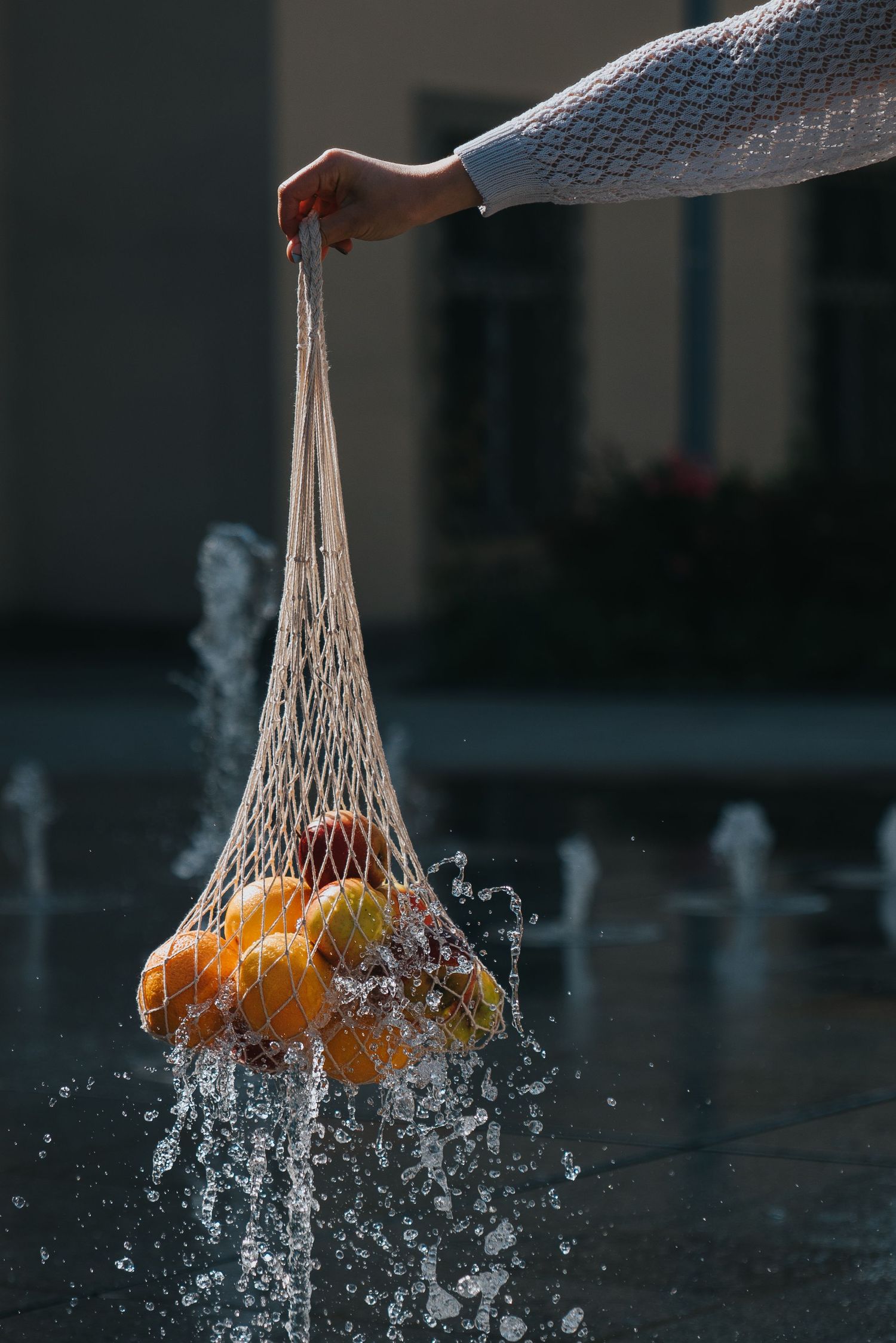 Hand holding a mesh string bag filled with fresh oranges being splashed with water near an outdoor fountain.