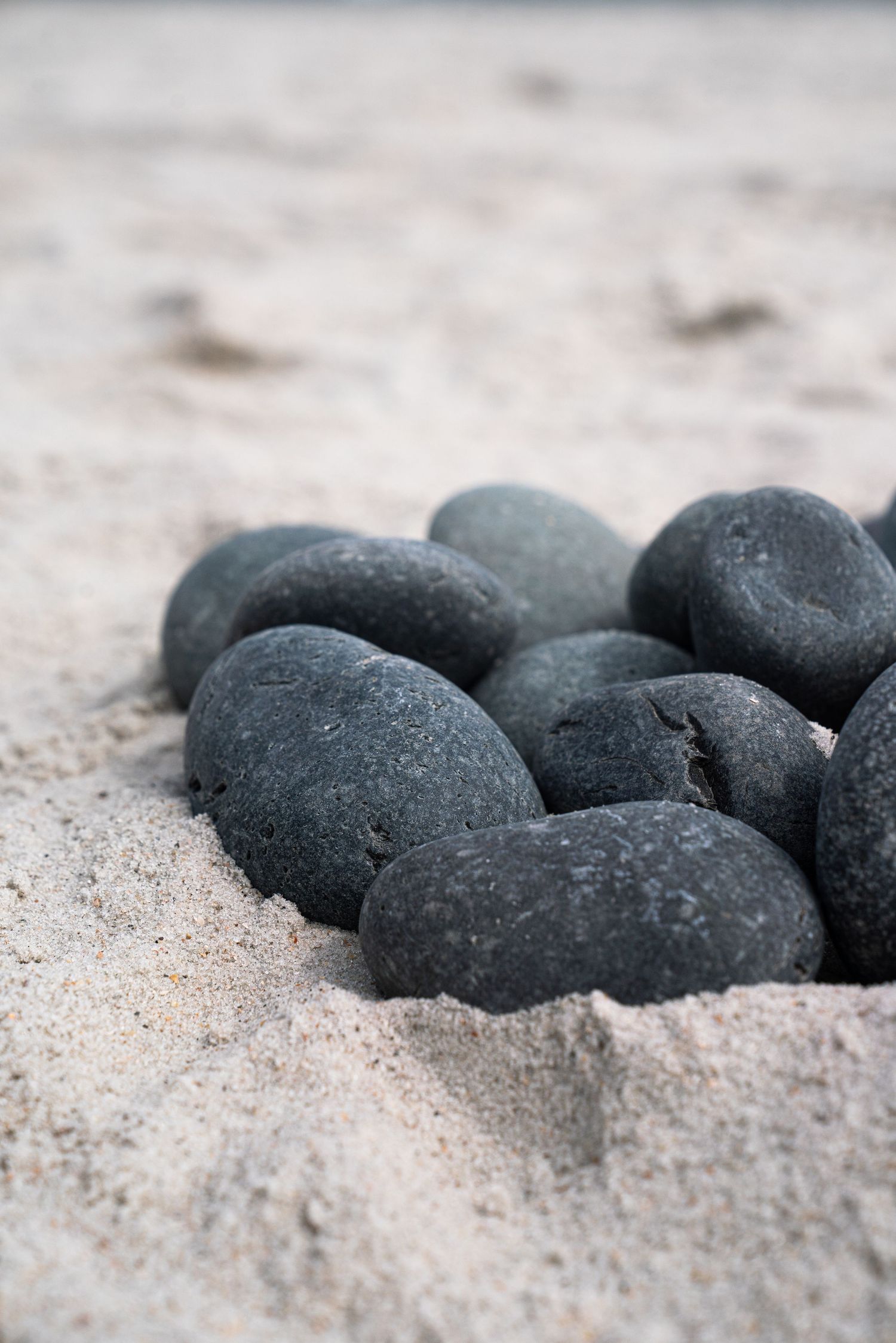 Dark gray smooth pebbles grouped together on light sandy beach surface in natural outdoor setting.