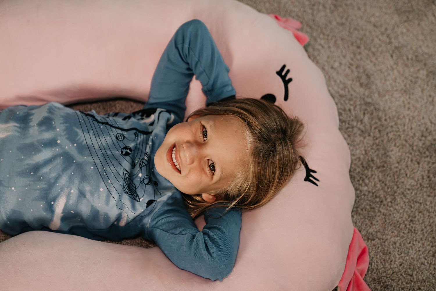 Smiling young girl lying on a pink unicorn floor cushion wearing a blue outfit, viewed from above.