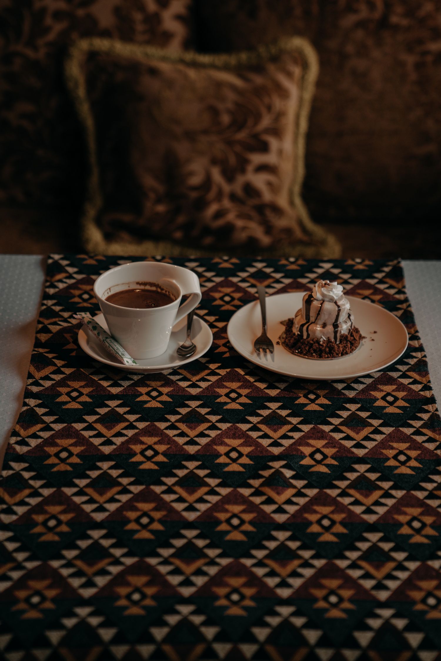 A cup of hot chocolate and a dessert plate on a patterned runner with decorative pillows in the background.