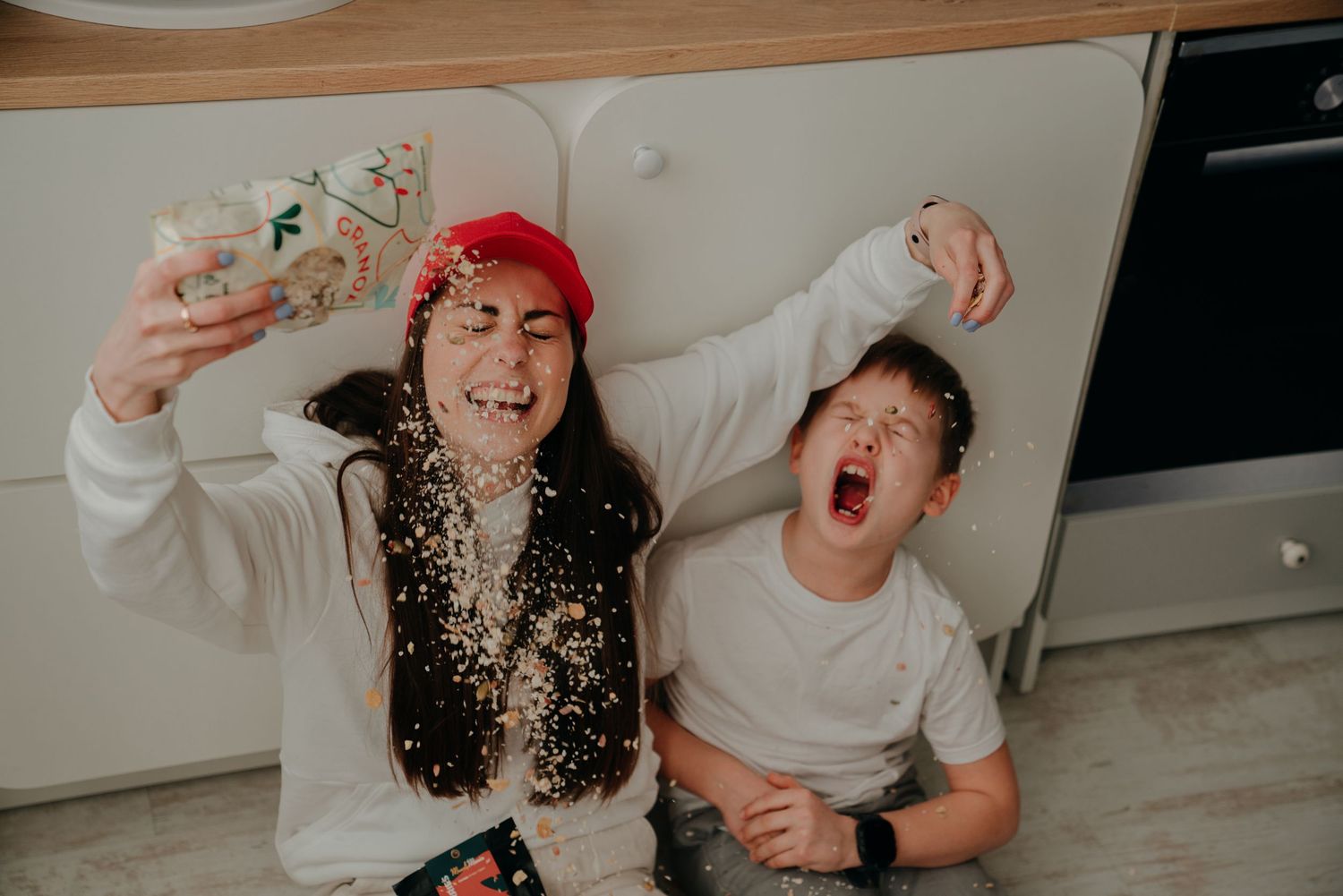 Two kids laughing and playing with glitter confetti on a kitchen floor, one wearing a red cap holding a jar.