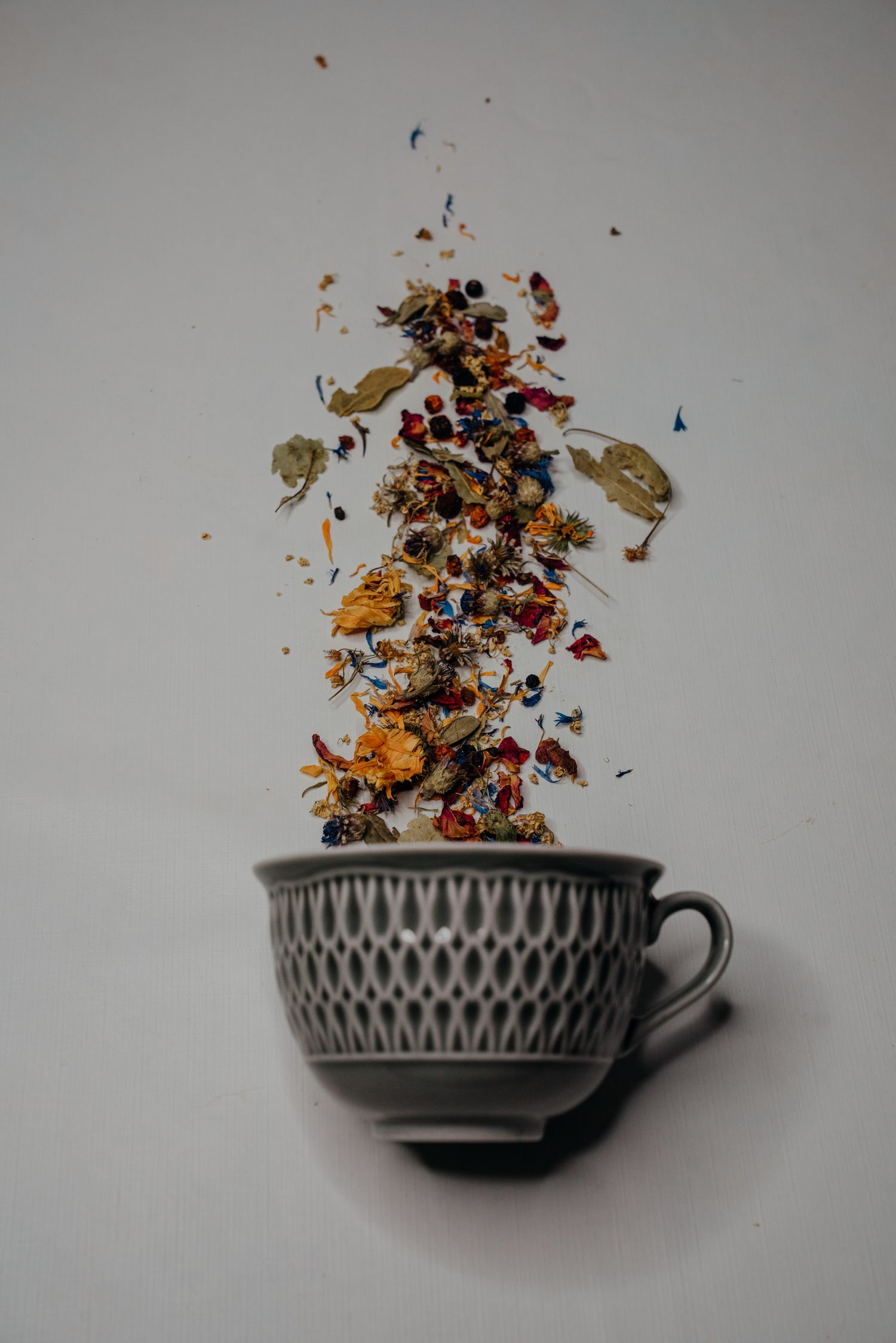 Dried herbal tea blend with colorful flowers and leaves floating above a gray ceramic cup on white background.