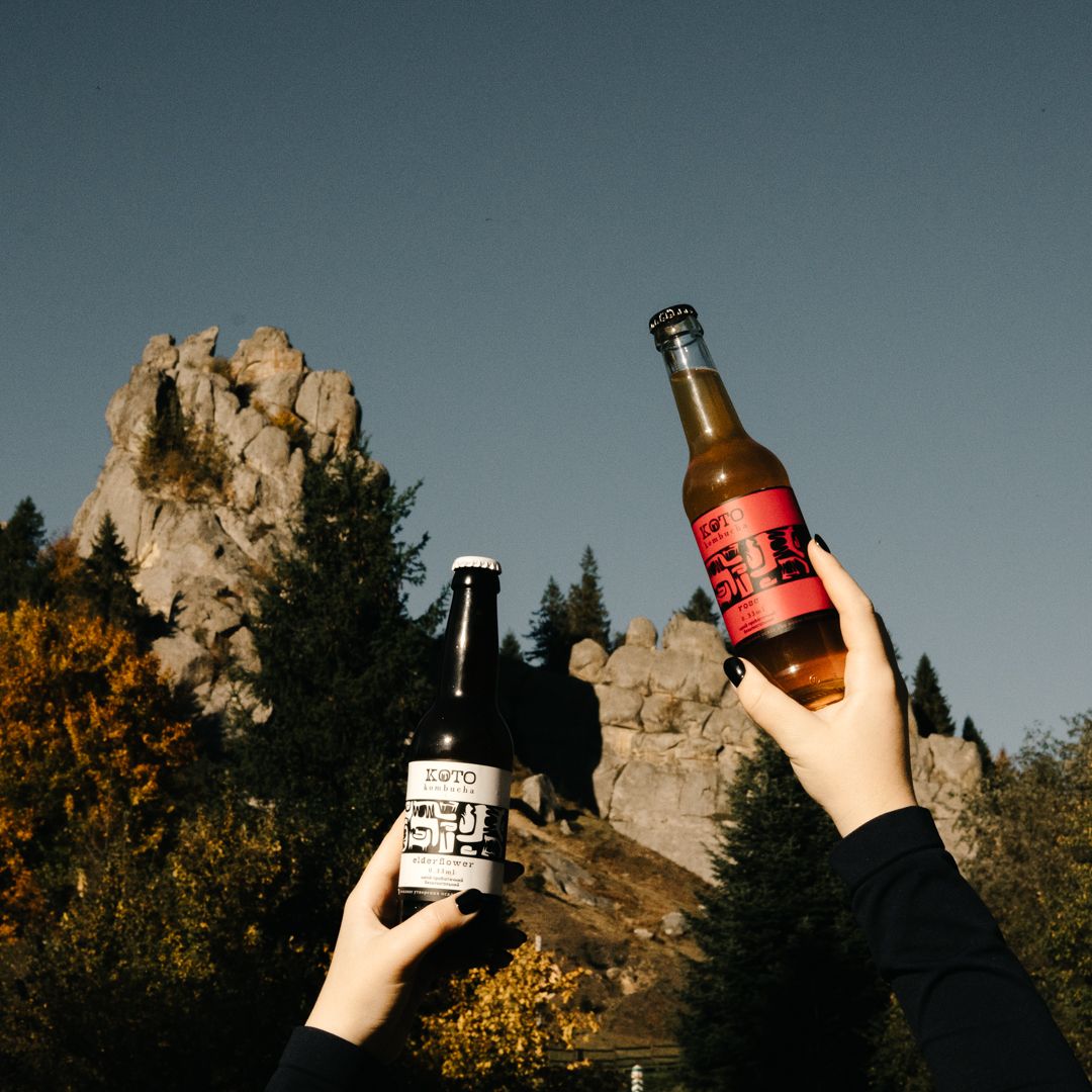 Two hands raising beer bottles against a dramatic rocky mountain landscape with autumn trees under a clear blue sky.