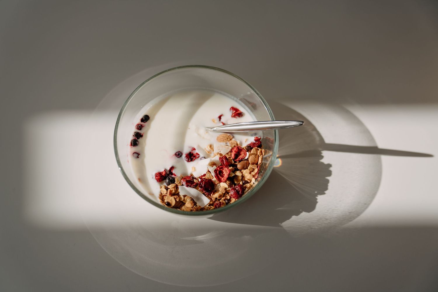 Glass bowl of creamy yogurt topped with granola and red berries, photographed from above in natural light.