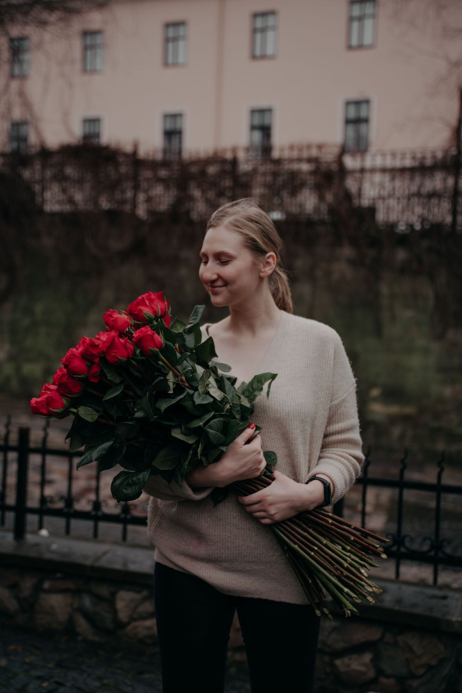 Smiling young woman holding a large bouquet of red roses outdoors near a black iron fence.