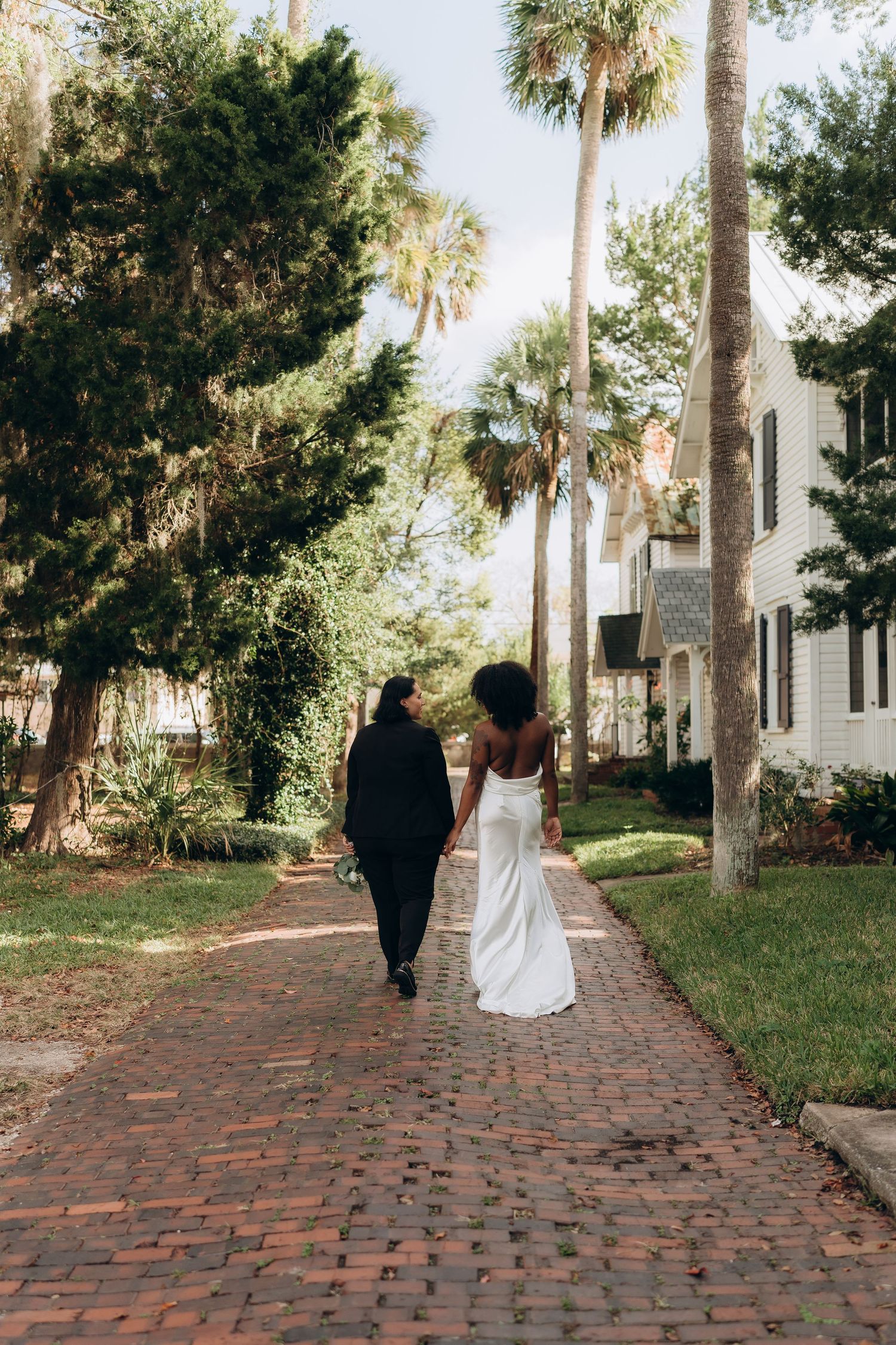 A couple walks along a palm tree-lined brick path in wedding attire.
