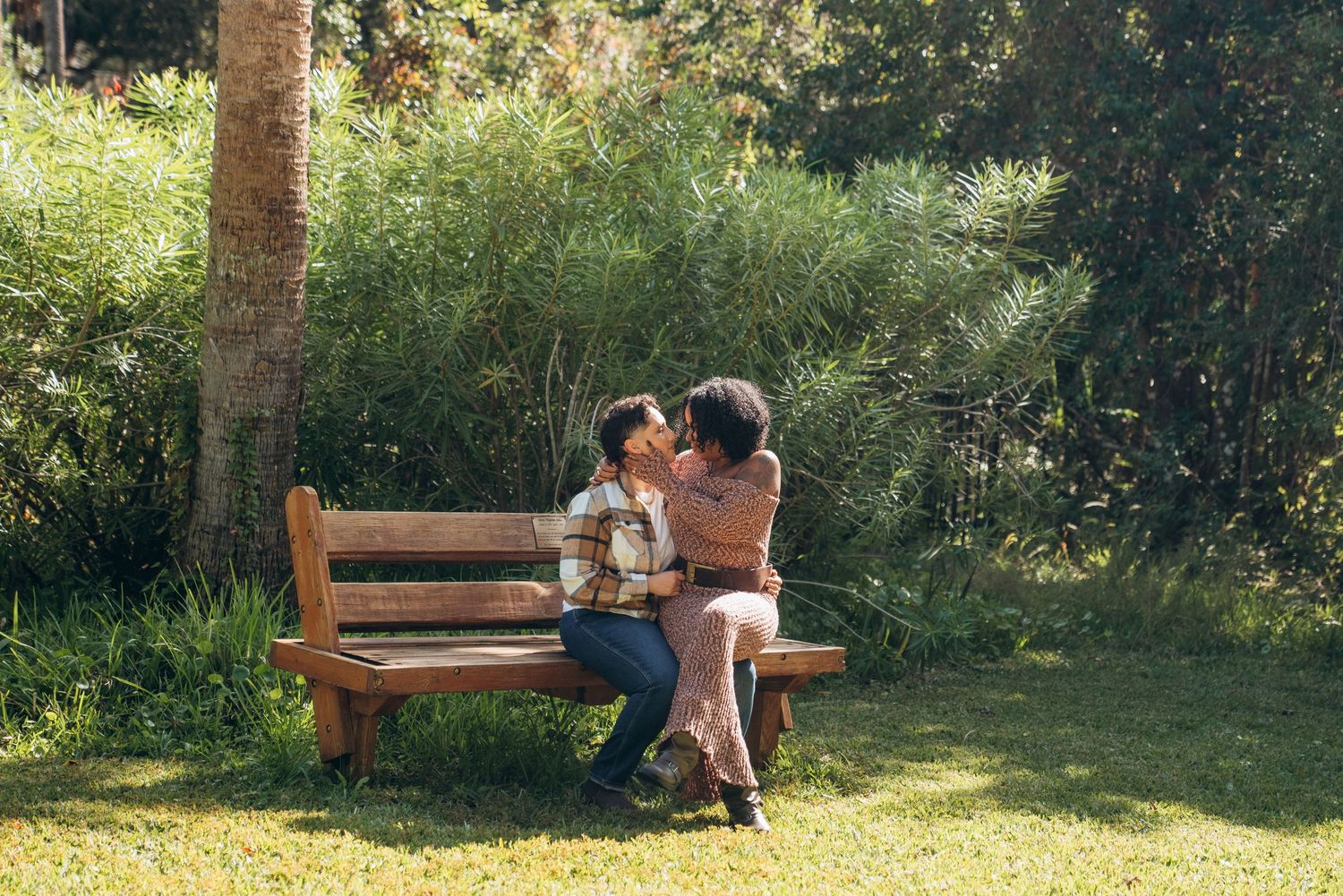 A couple sits on a wooden bench in a sunny garden, sharing an intimate moment surrounded by lush greenery.