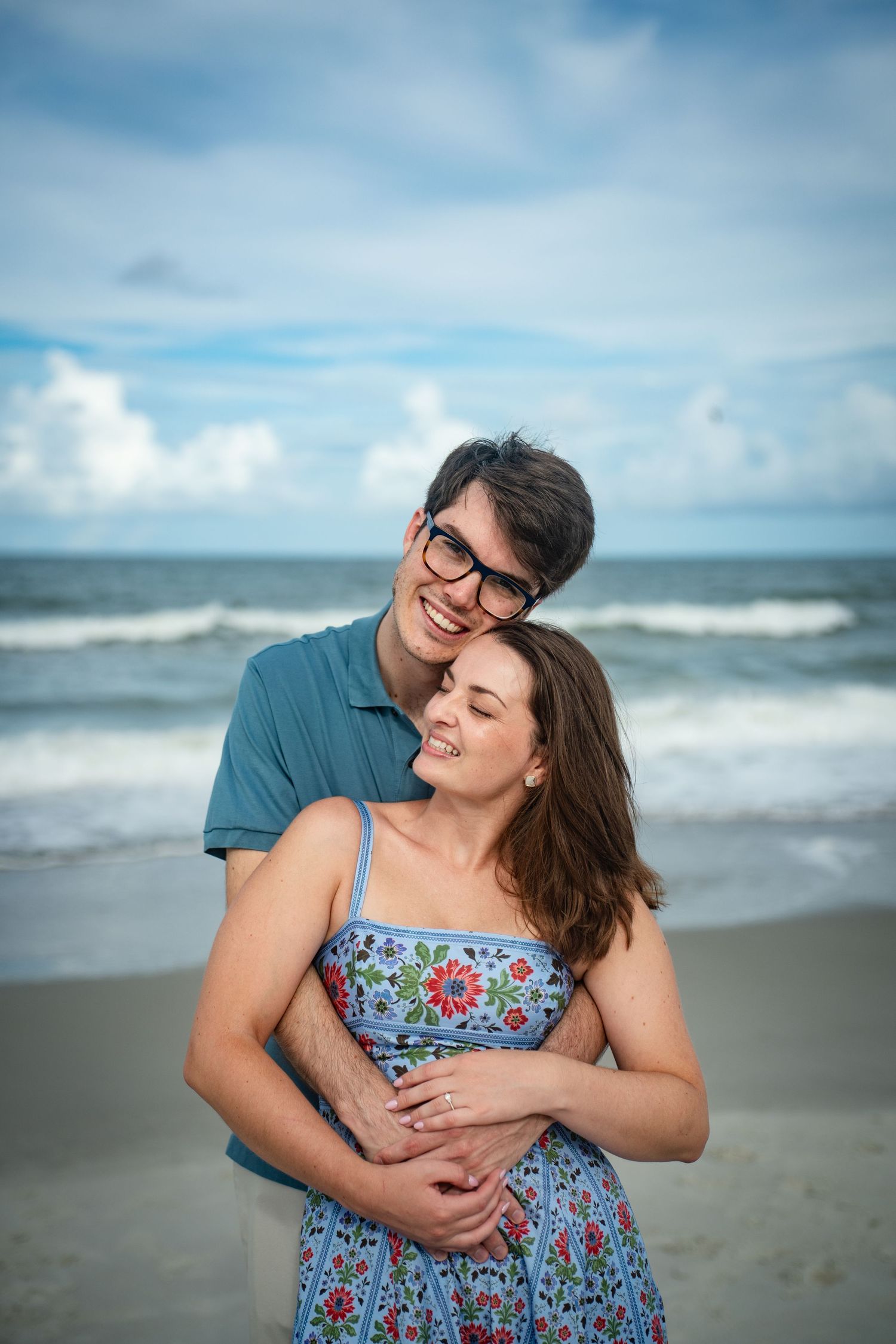 A couple embraces joyfully on a cloudy beach, with the woman wearing a floral blue dress and waves in the background.
