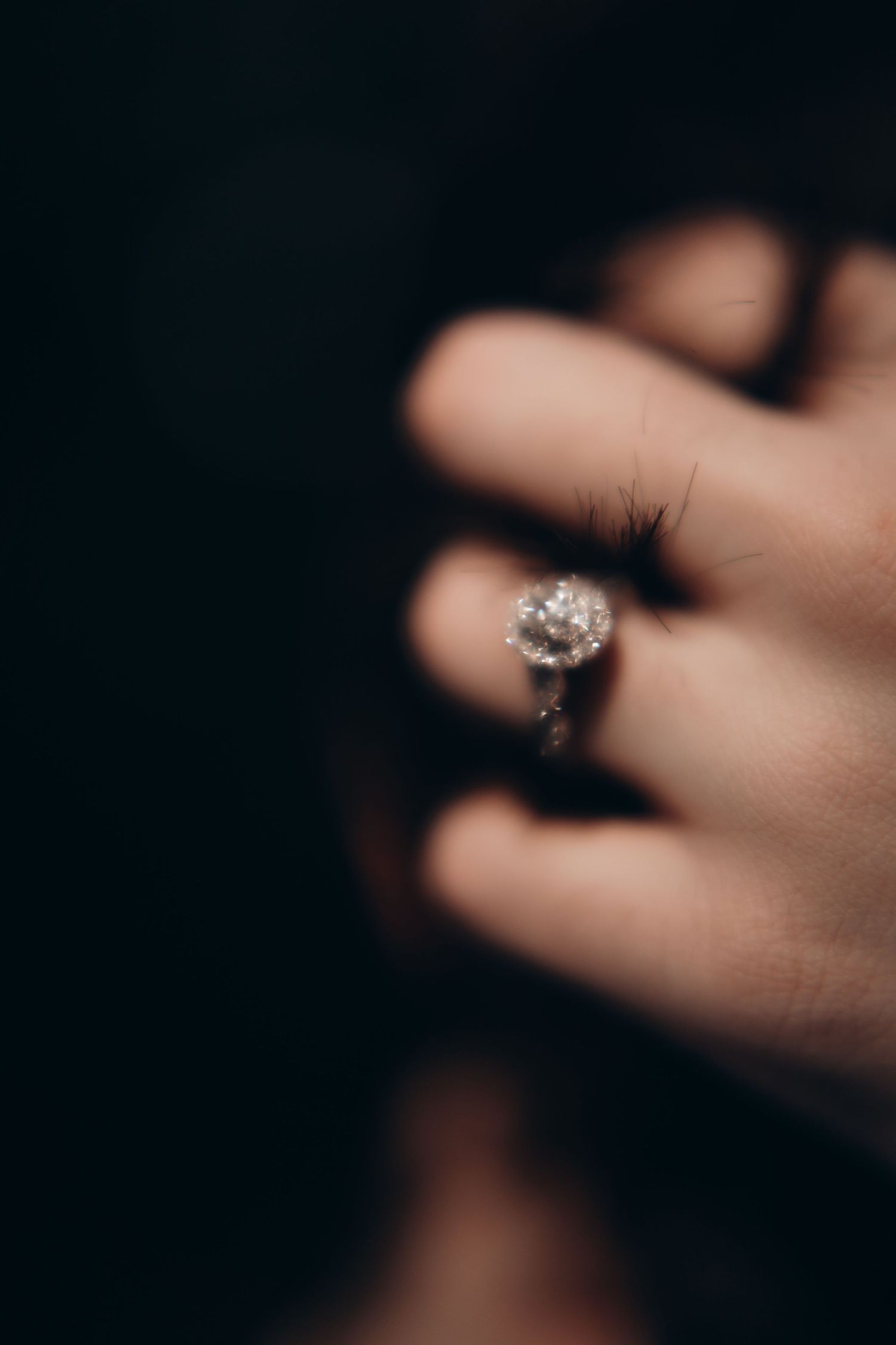 Close-up view of a sparkling diamond engagement ring on a hand against a dark background.