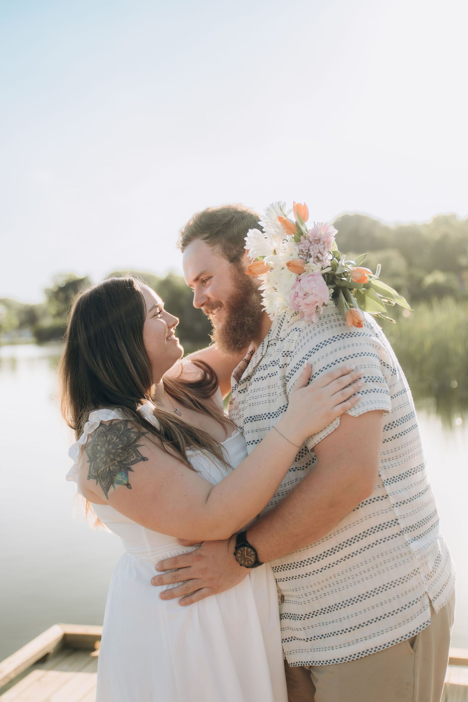 A romantic couple embraces by a lake at sunset, with soft lighting and floral decorations creating a dreamy atmosphere.