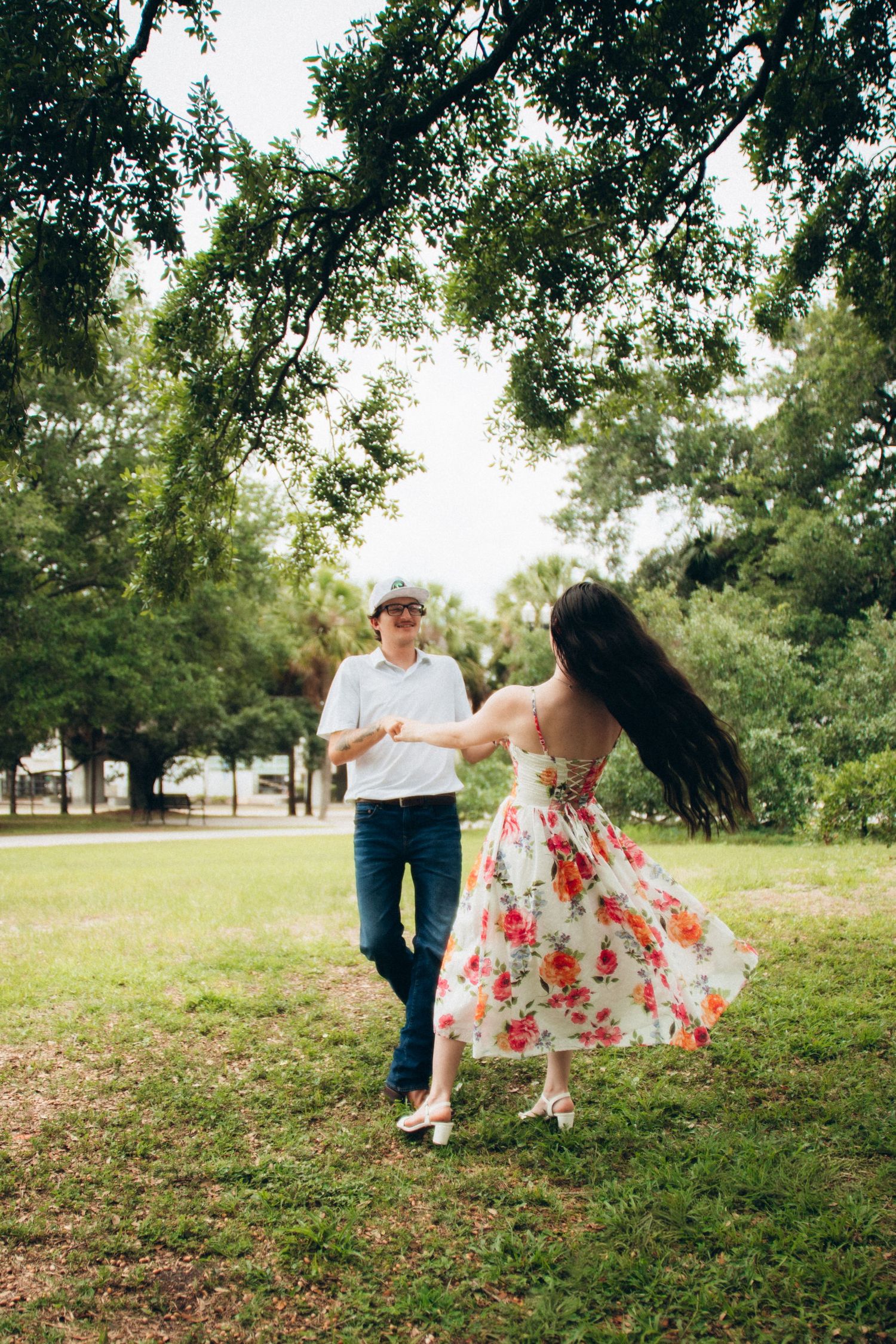 Couple dancing together in park as woman in floral dress twirls holding mans hands under trees.