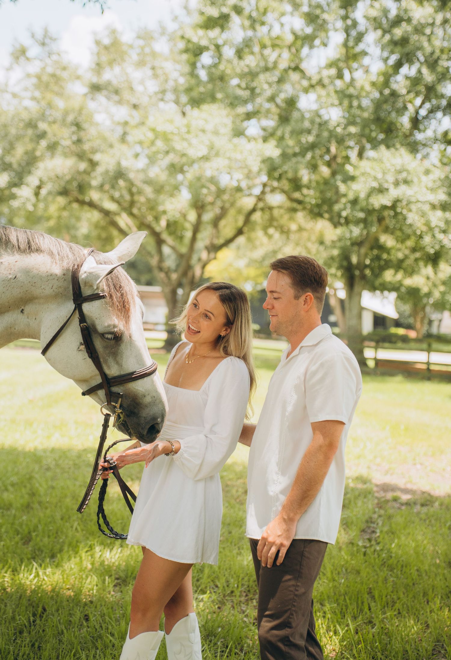A couple shares a playful moment with their white horse in a sunny pasture.