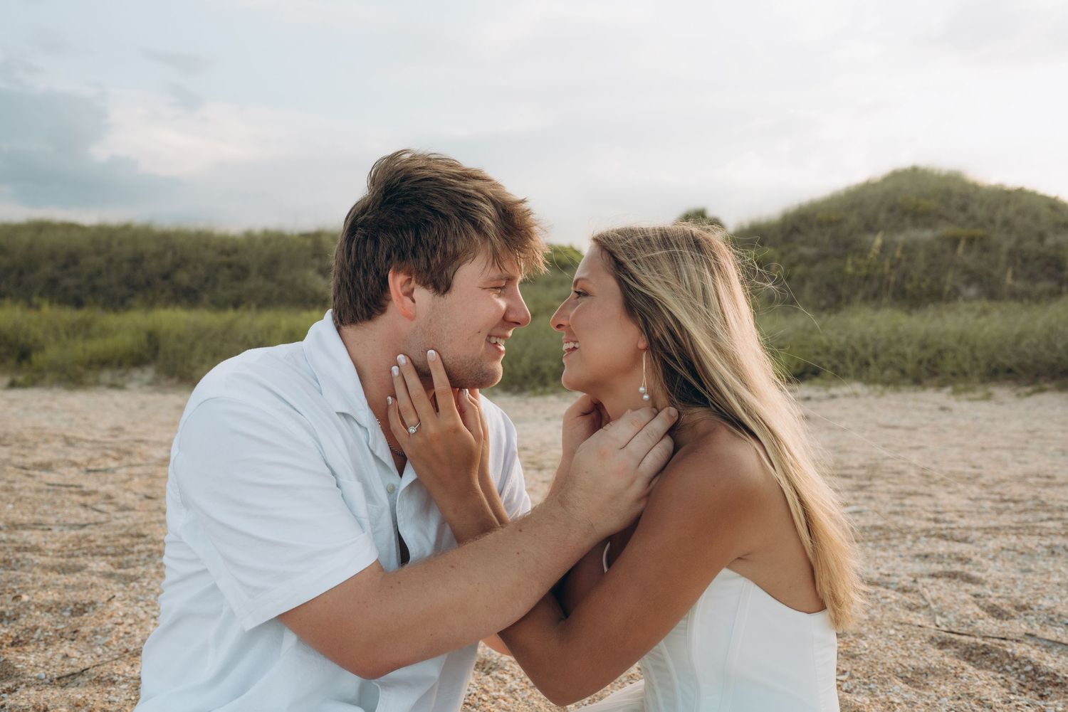 Intimate beach photoshoot with couple in white outfits against coastal backdrop.