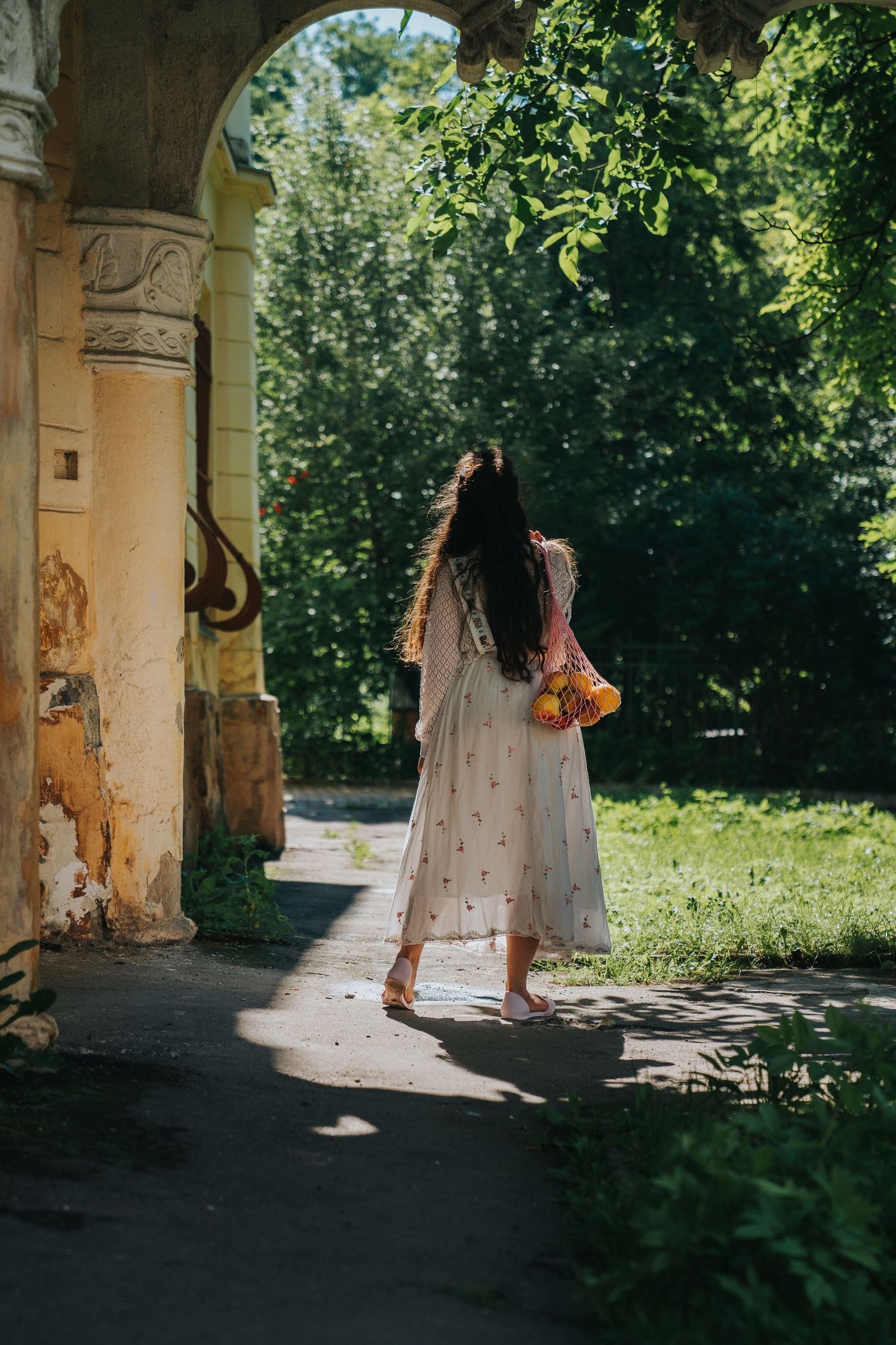 Woman in white floral dress carrying yellow flowers walking through a sunlit garden archway.