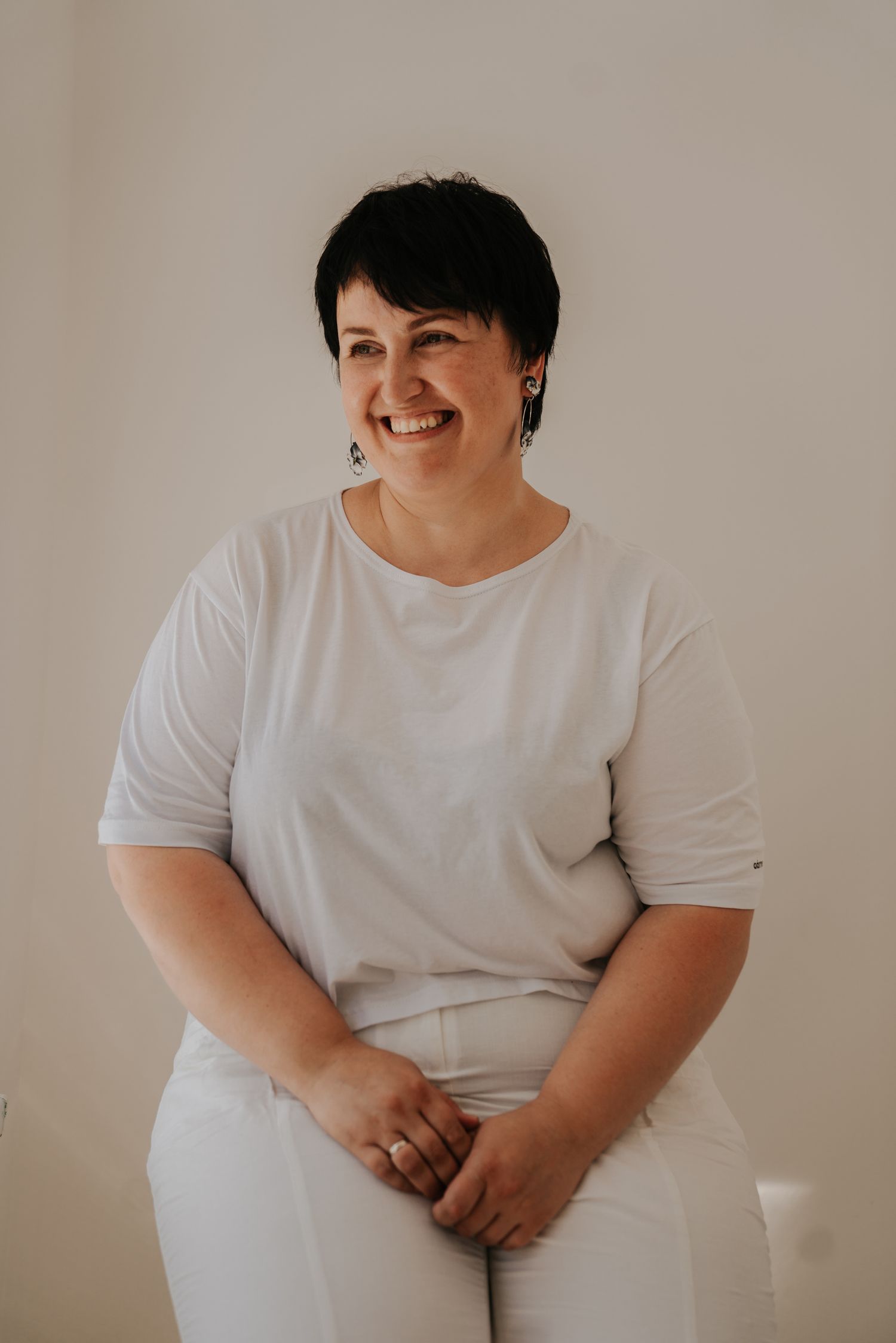 Smiling woman with short dark hair wearing white t-shirt and pants, sitting relaxed against a light background.