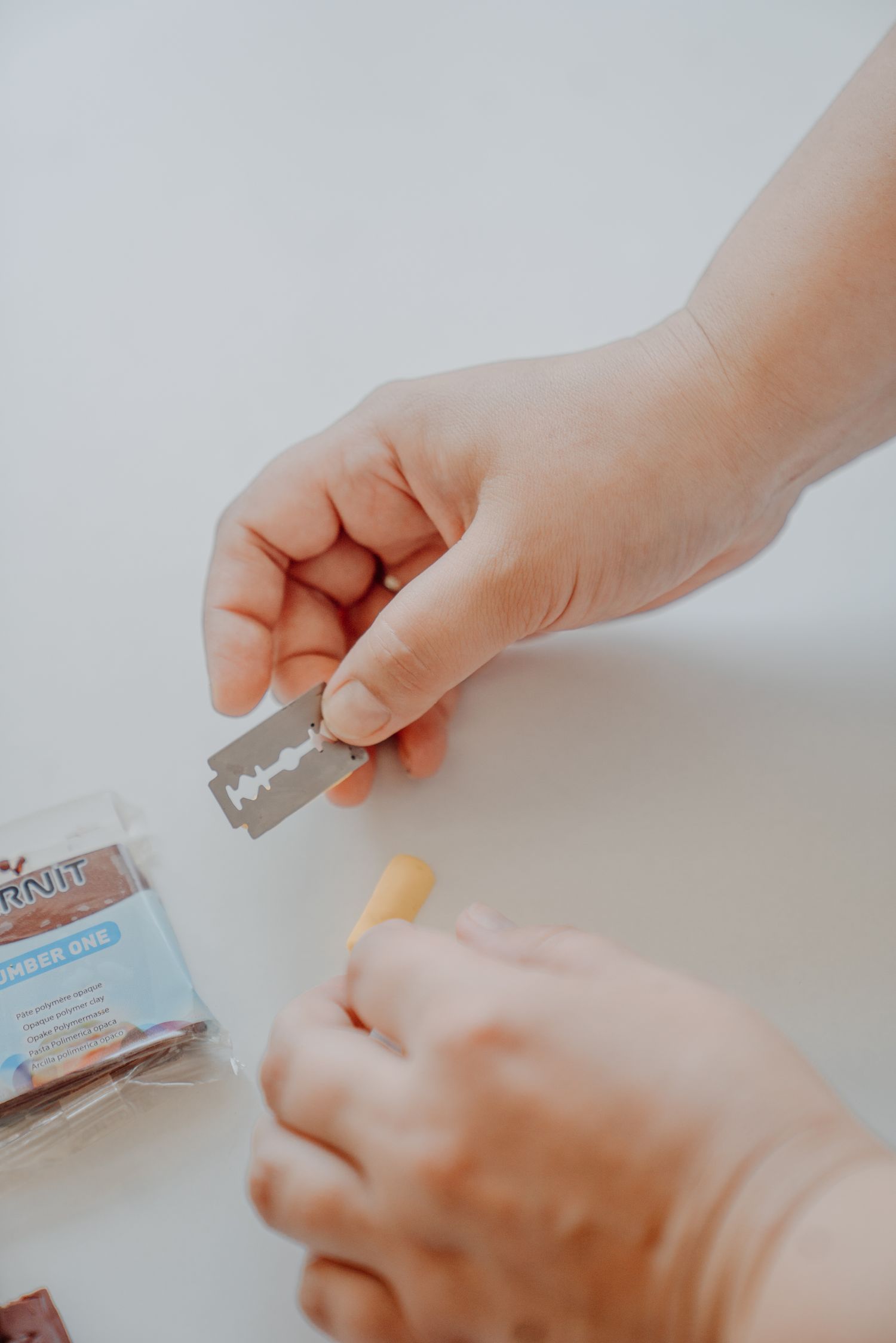 Hands holding a razor blade and small piece of material on a white surface with craft supplies nearby.