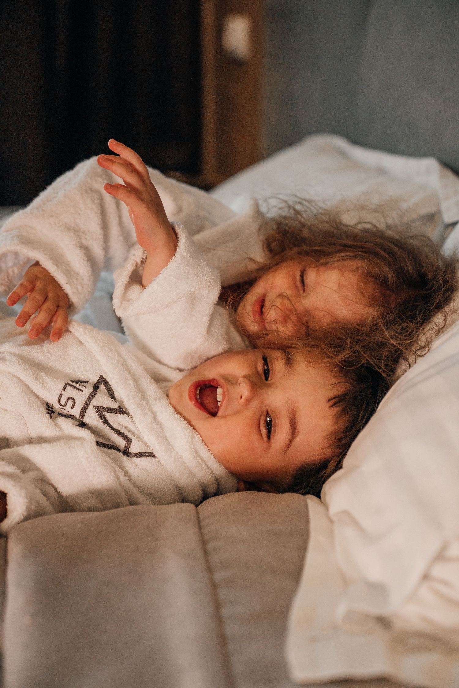 A young child in a white hotel robe laughing and playing on a bed with white linens.