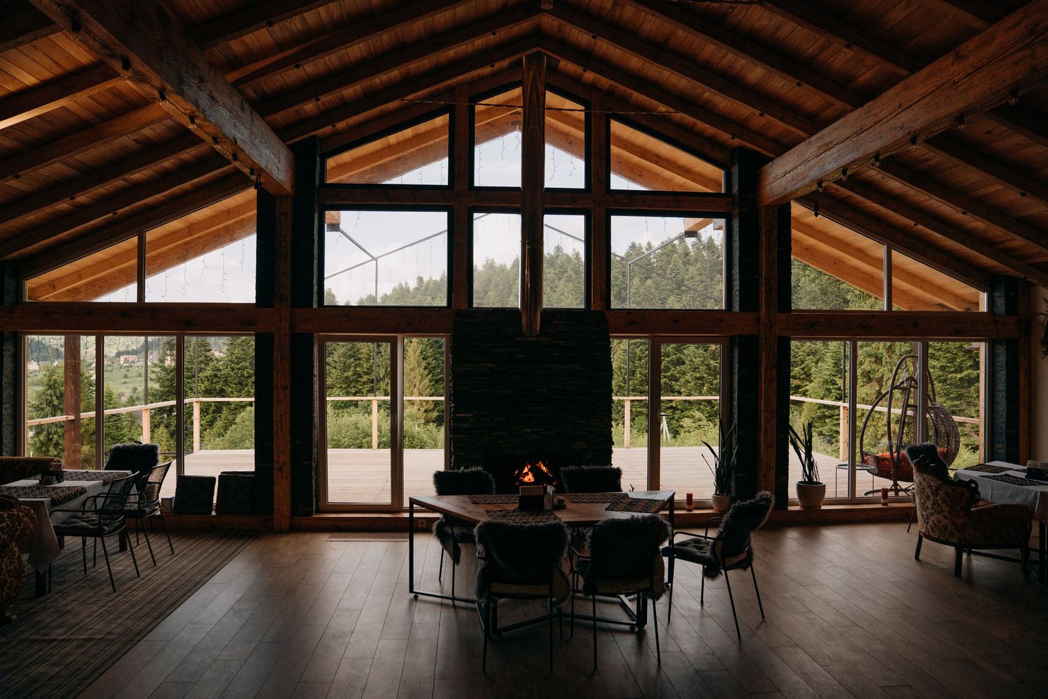 Rustic cabin interior with vaulted wood ceiling, fireplace, dining area, and floor-to-ceiling windows overlooking pine forest.