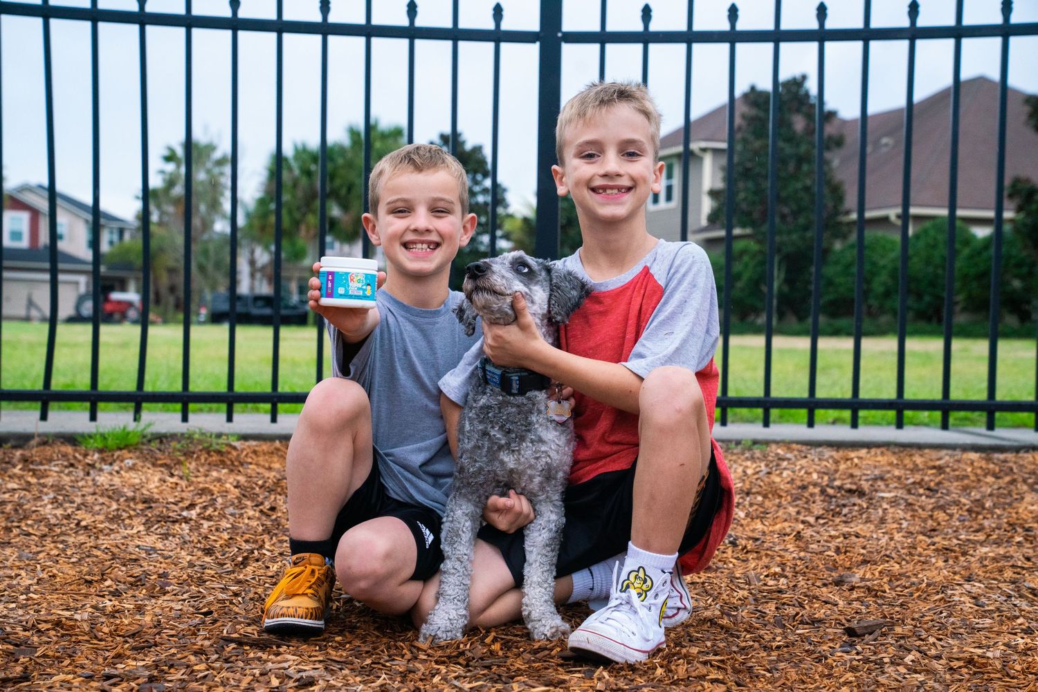 Two smiling boys crouching with a gray dog holding a supplement jar outdoors.