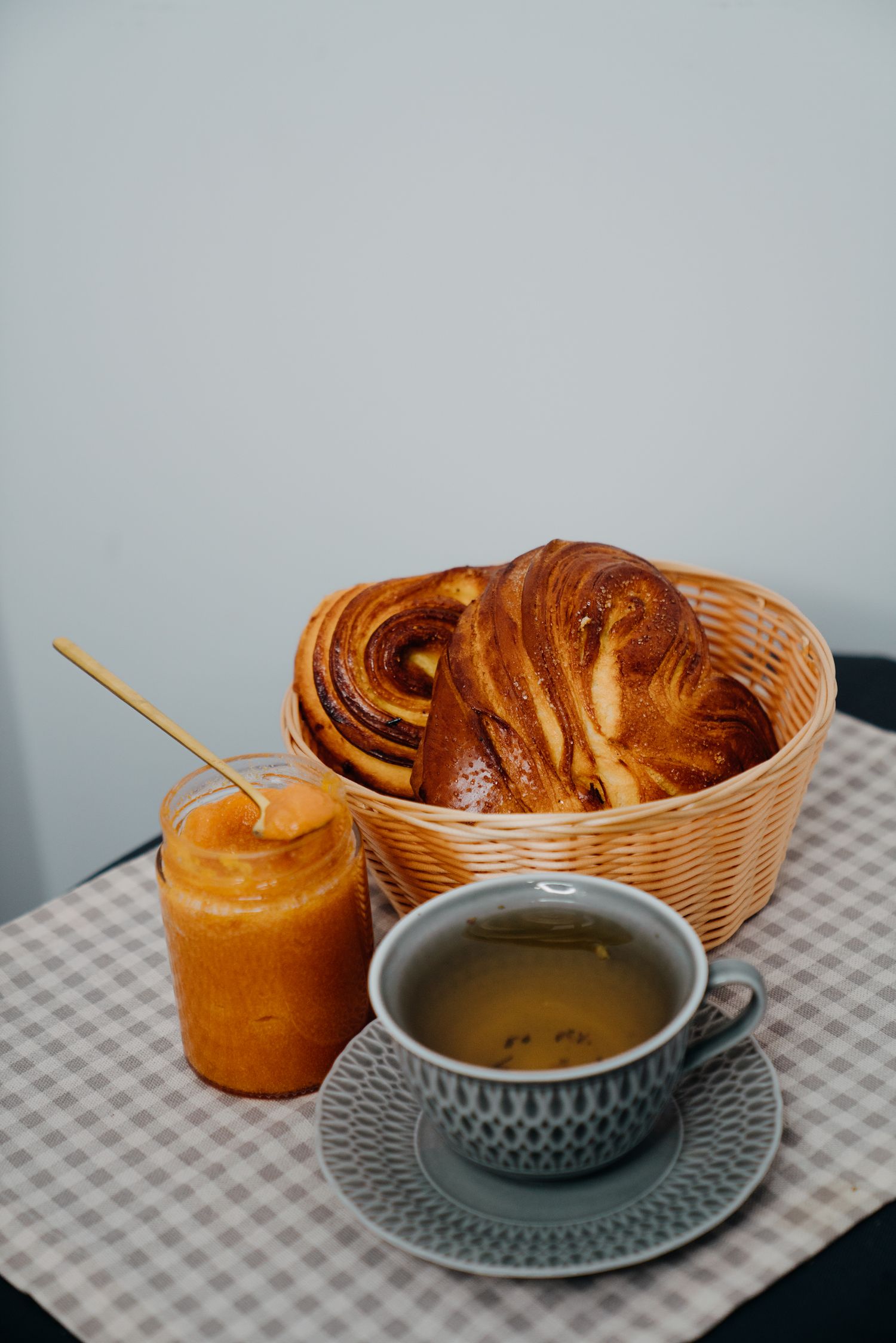 Wicker basket with golden pastries beside a jar of apricot jam and a cup of green tea on checkered tablecloth.