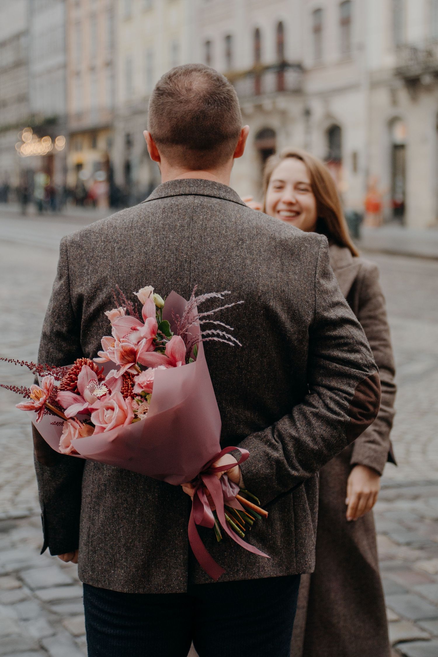 Man hiding pink flower bouquet behind his back to surprise smiling woman on a cobblestone city street.