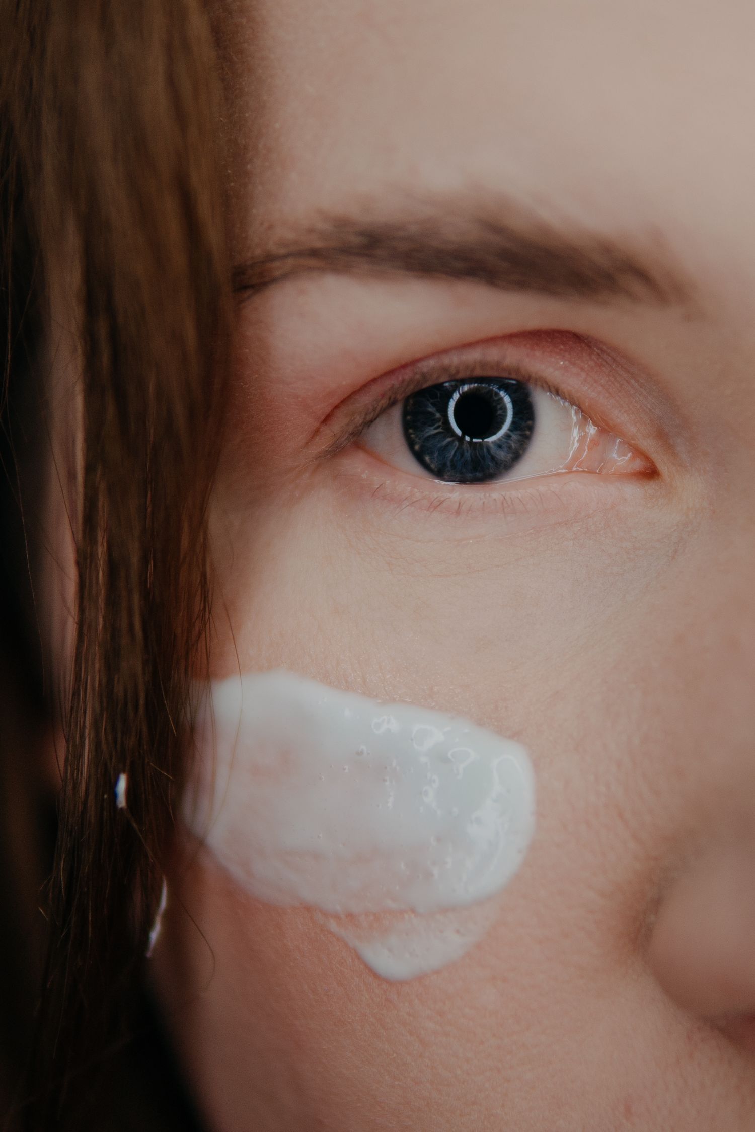 Close-up of a woman with blue eyes applying white face cream to her cheek for skincare routine.