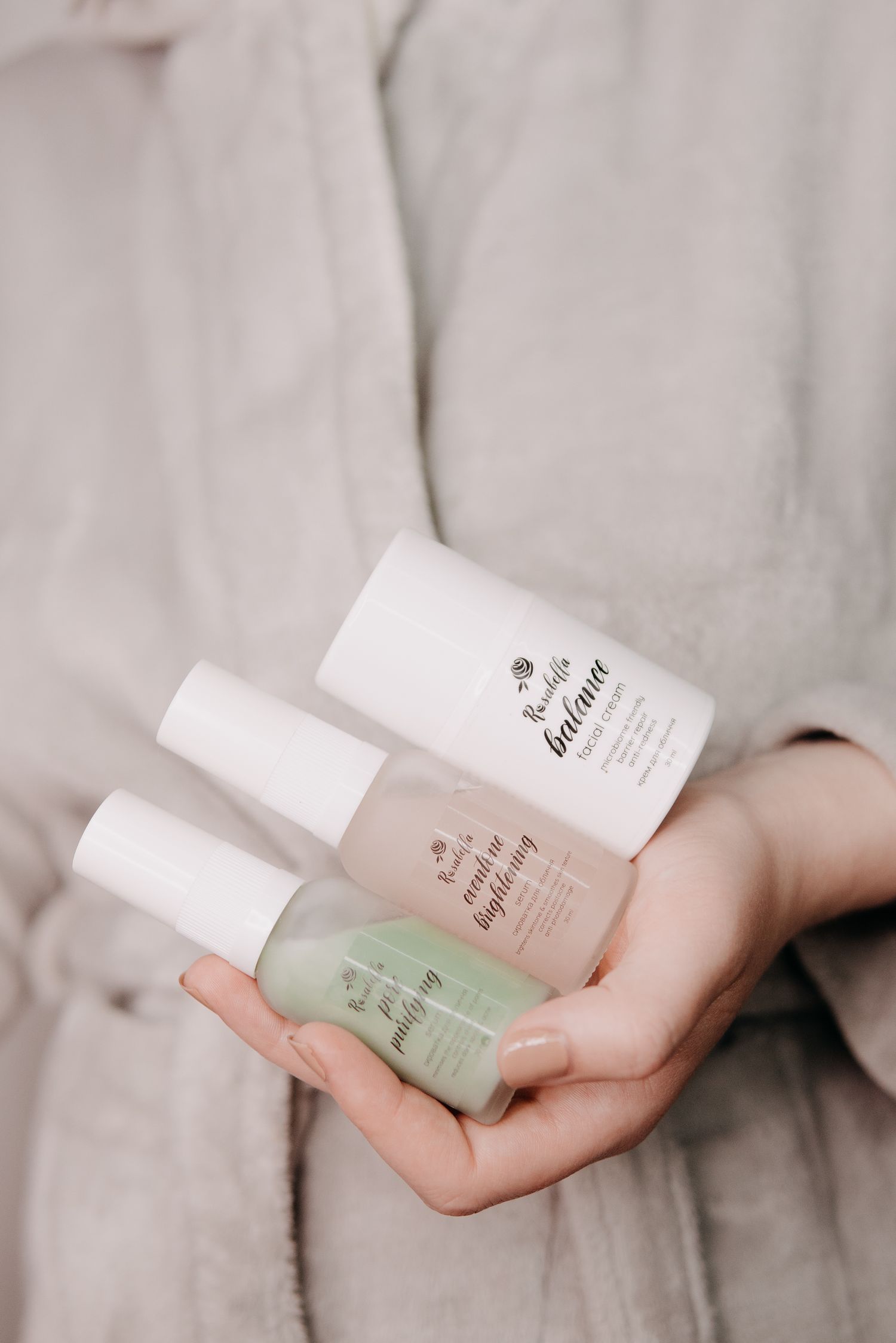 Hand holding three skincare bottles, including balance and purifying serums, against a soft linen background.