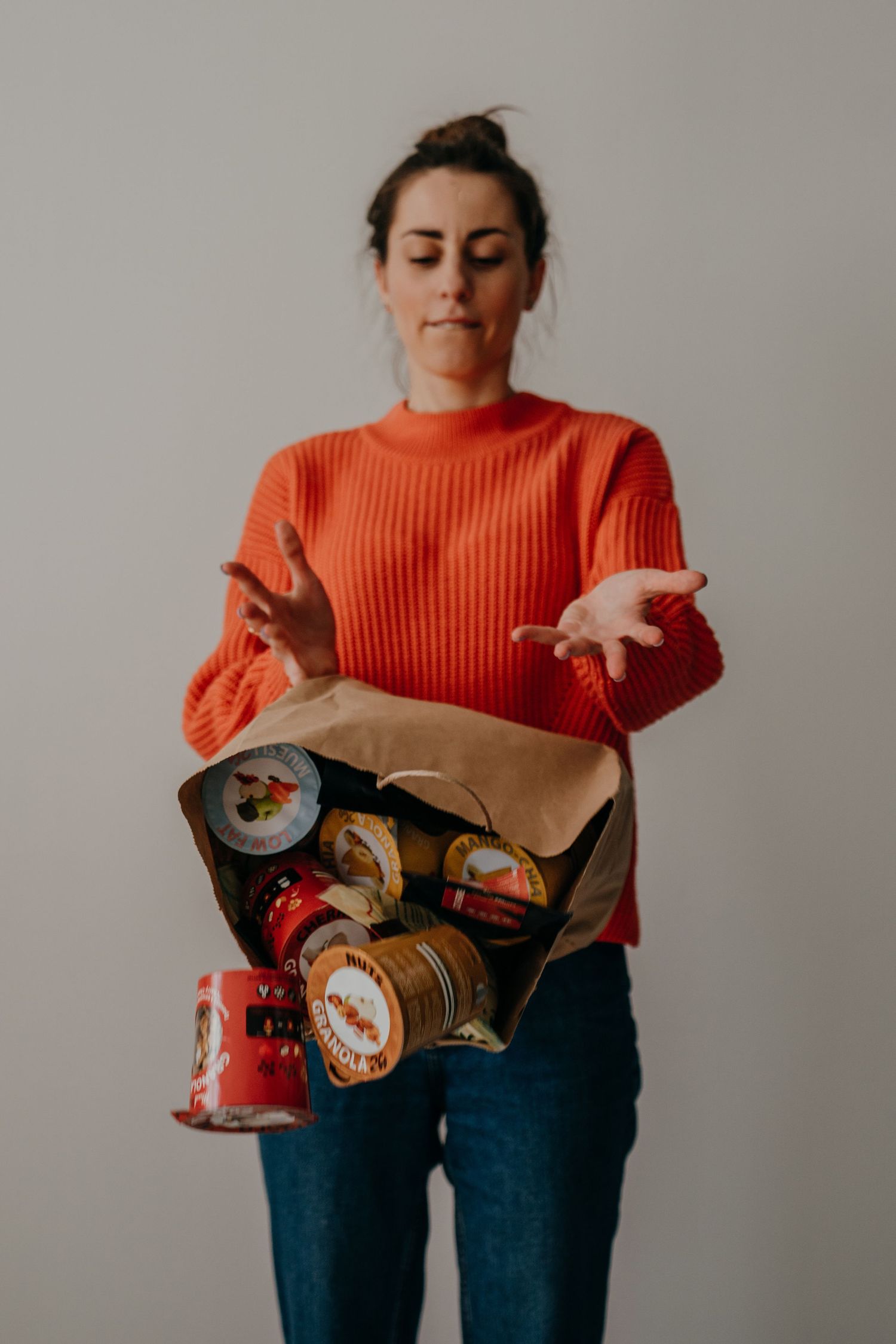Woman in orange sweater holding a paper bag filled with canned goods, looking puzzled with hands gesturing outward.