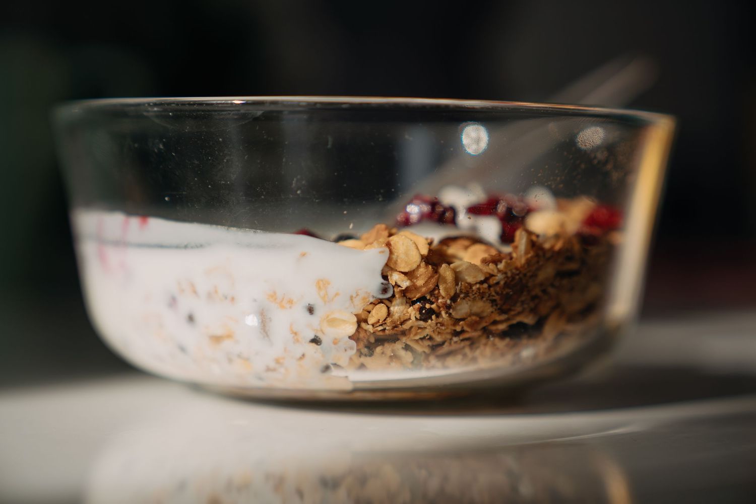 Close-up of a glass bowl filled with granola, yogurt, and red berries on a reflective surface.