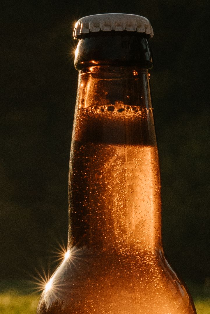 Close-up of a cold amber beer bottle with condensation, backlit by warm golden sunlight with a white bottle cap.