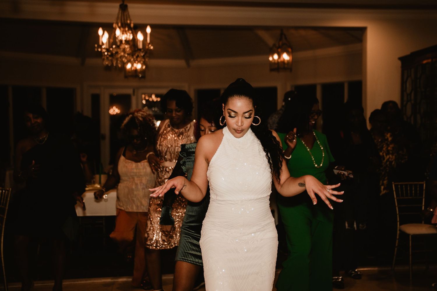 Bride in white halter gown dancing joyfully at wedding reception with guests in elegant attire under chandelier lighting.