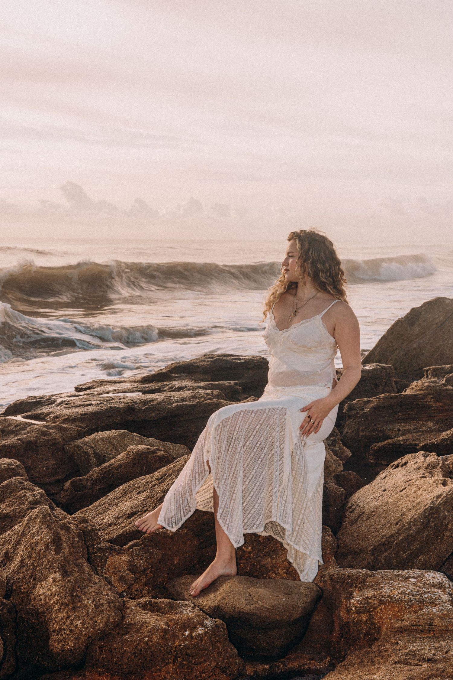 Woman in white lace dress sitting on rocky shore with ocean waves crashing behind her at sunset.