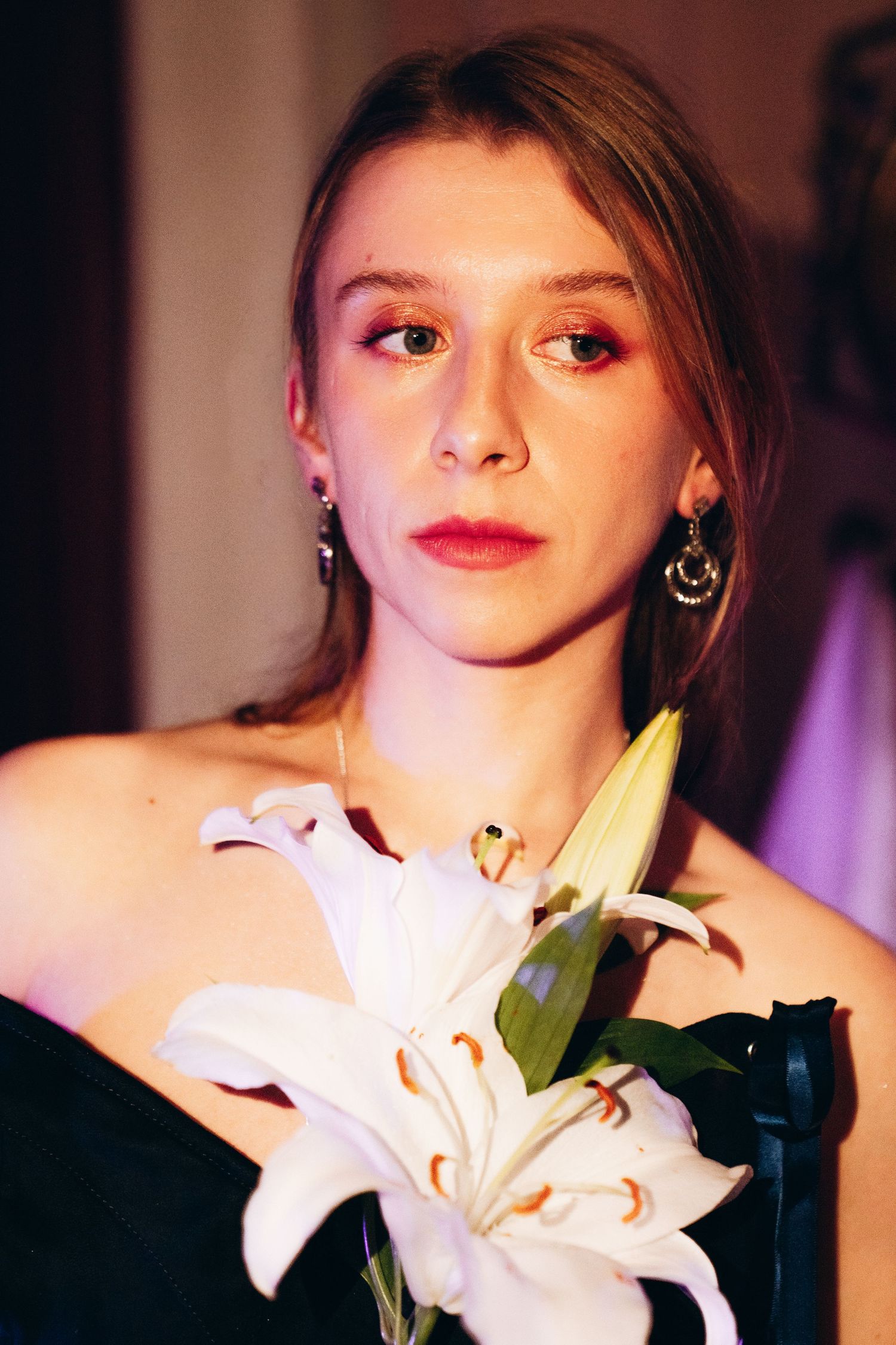 Close-up portrait of young woman with pink eye makeup holding white lily flower wearing black off-shoulder top.