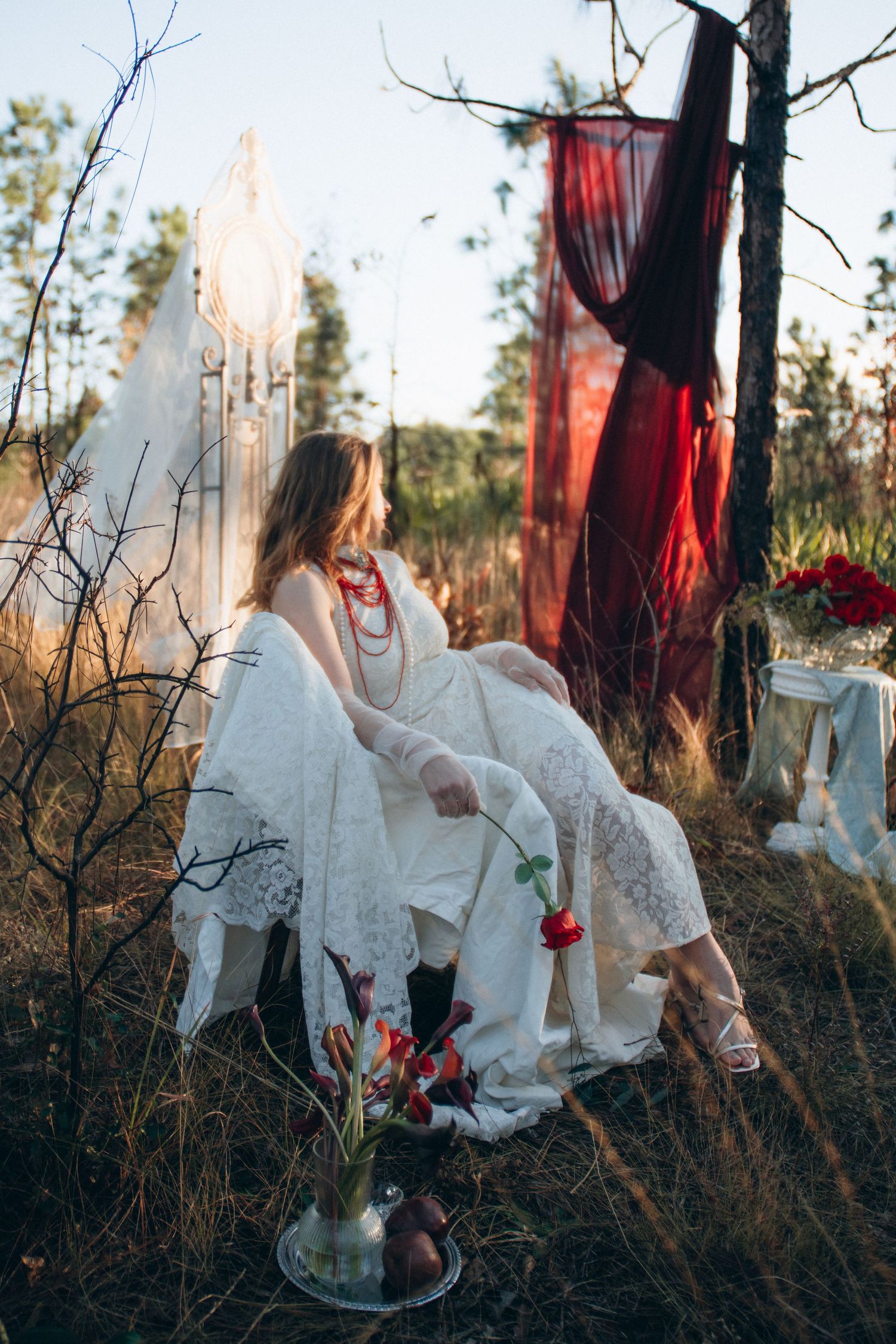 A bohemian bride in white lace dress sits outdoors among wildgrass with red fabric draping and floral arrangements nearby.